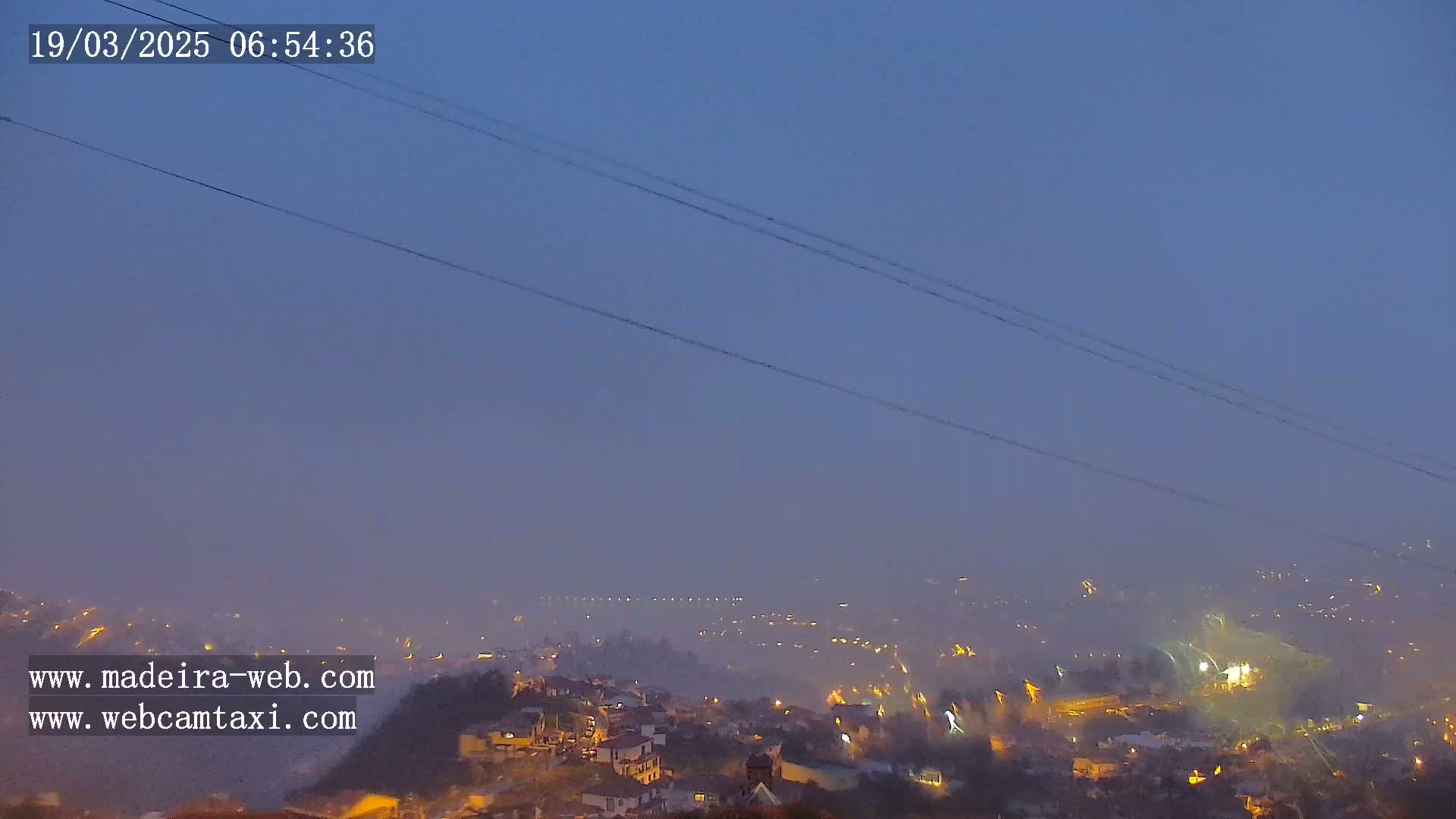 A hazy, dimly lit town is seen from a distance at twilight, with power lines crossing the frame.