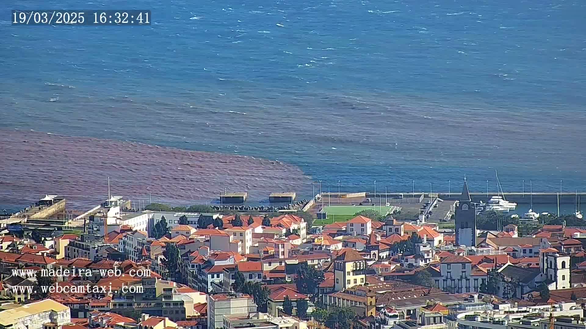A coastal town with red-tinged water meeting blue ocean water under a clear sky.