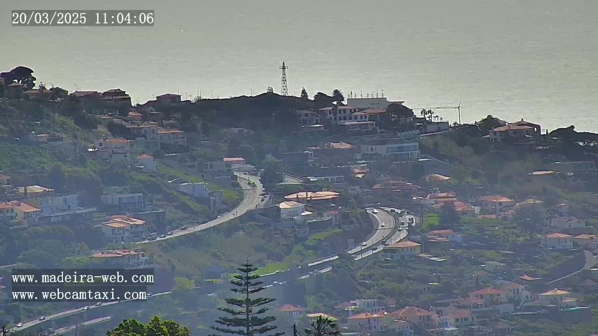 A hazy, coastal hillside town with a winding road and numerous houses overlooks a calm ocean.