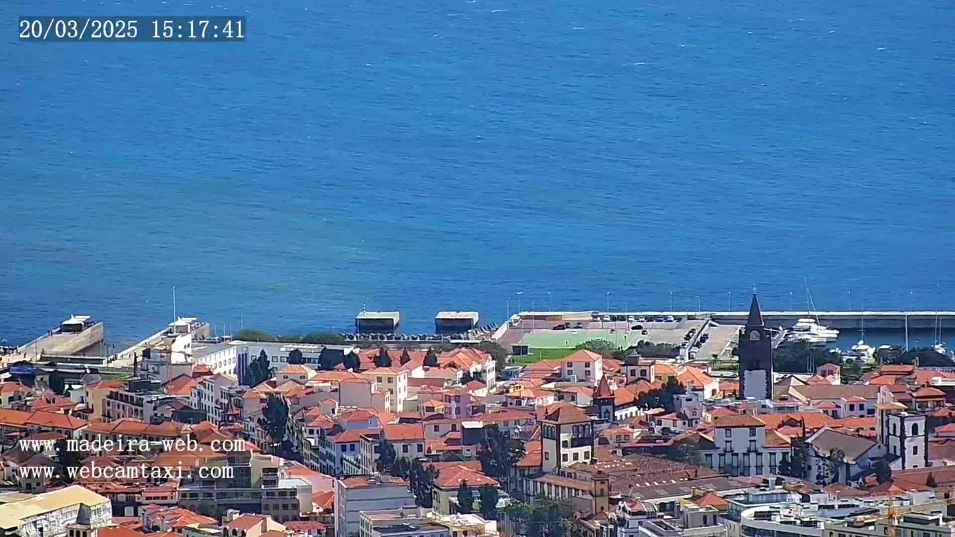 A coastal town with red-roofed buildings and a tall, dark clock tower is situated next to a calm, blue ocean on a sunny day.