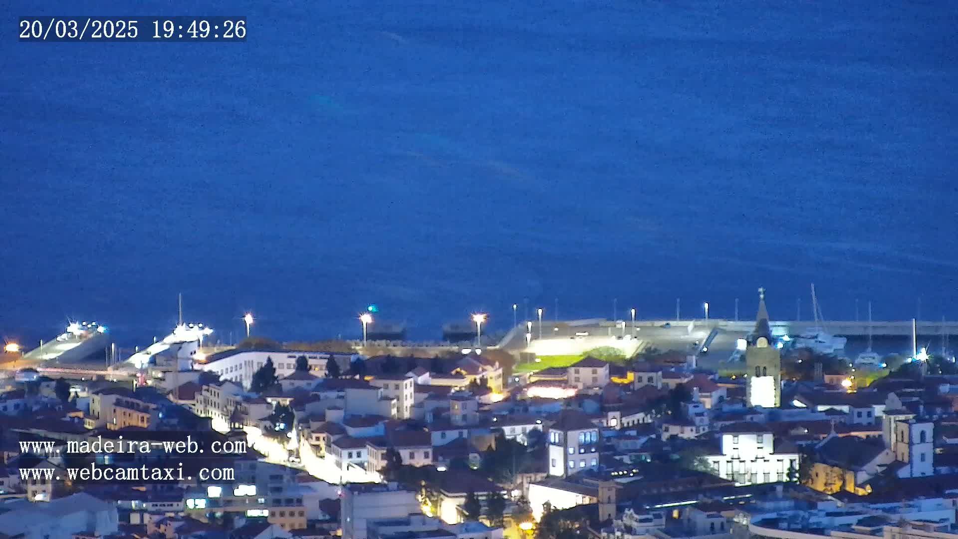 A coastal town at night, illuminated with lights, is situated next to a dark blue body of water under a clear night sky.