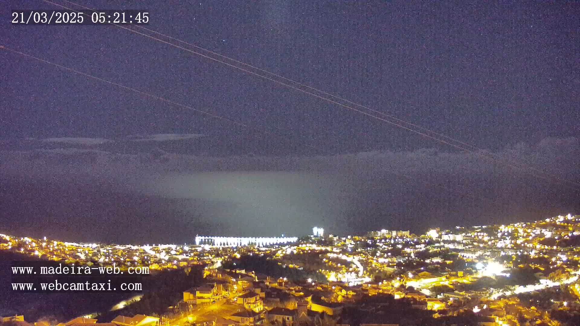 A nighttime view of a city illuminated by lights under a mostly clear, dark sky with a few clouds and faint stars.