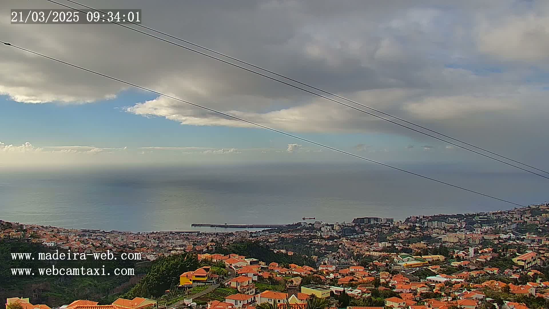 A coastal city with red-tiled roofs is visible below a partly cloudy sky, overlooking a calm ocean.