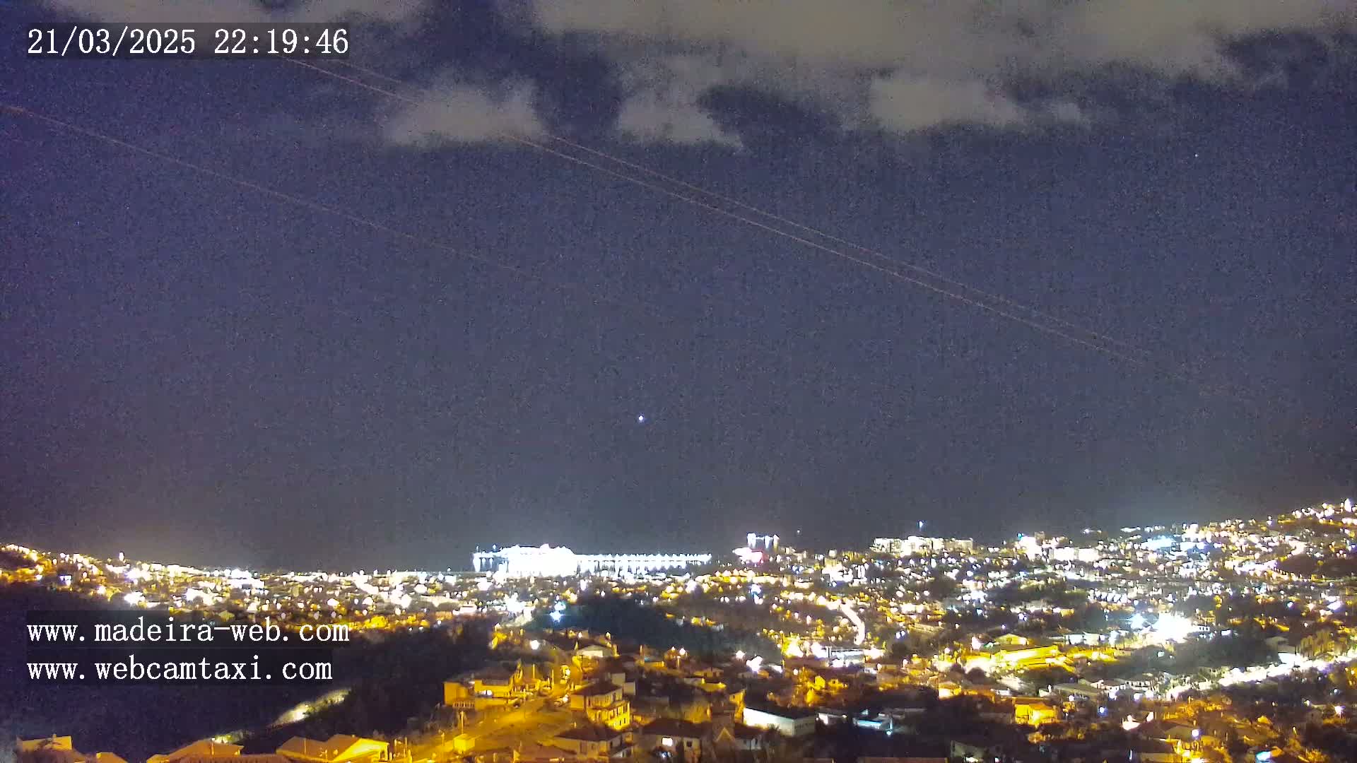 A nighttime view of a city illuminated by lights under a partly cloudy sky shows two airplane contrails.