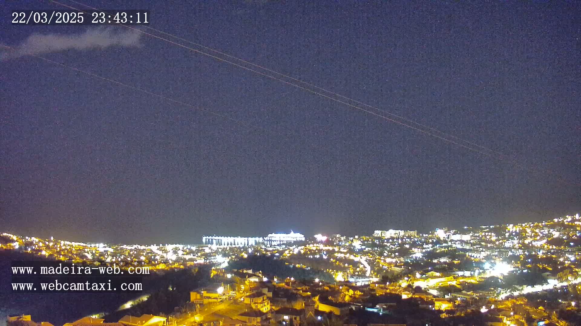 A nighttime view of a city illuminated by lights under a mostly clear, dark sky with a few faint clouds.