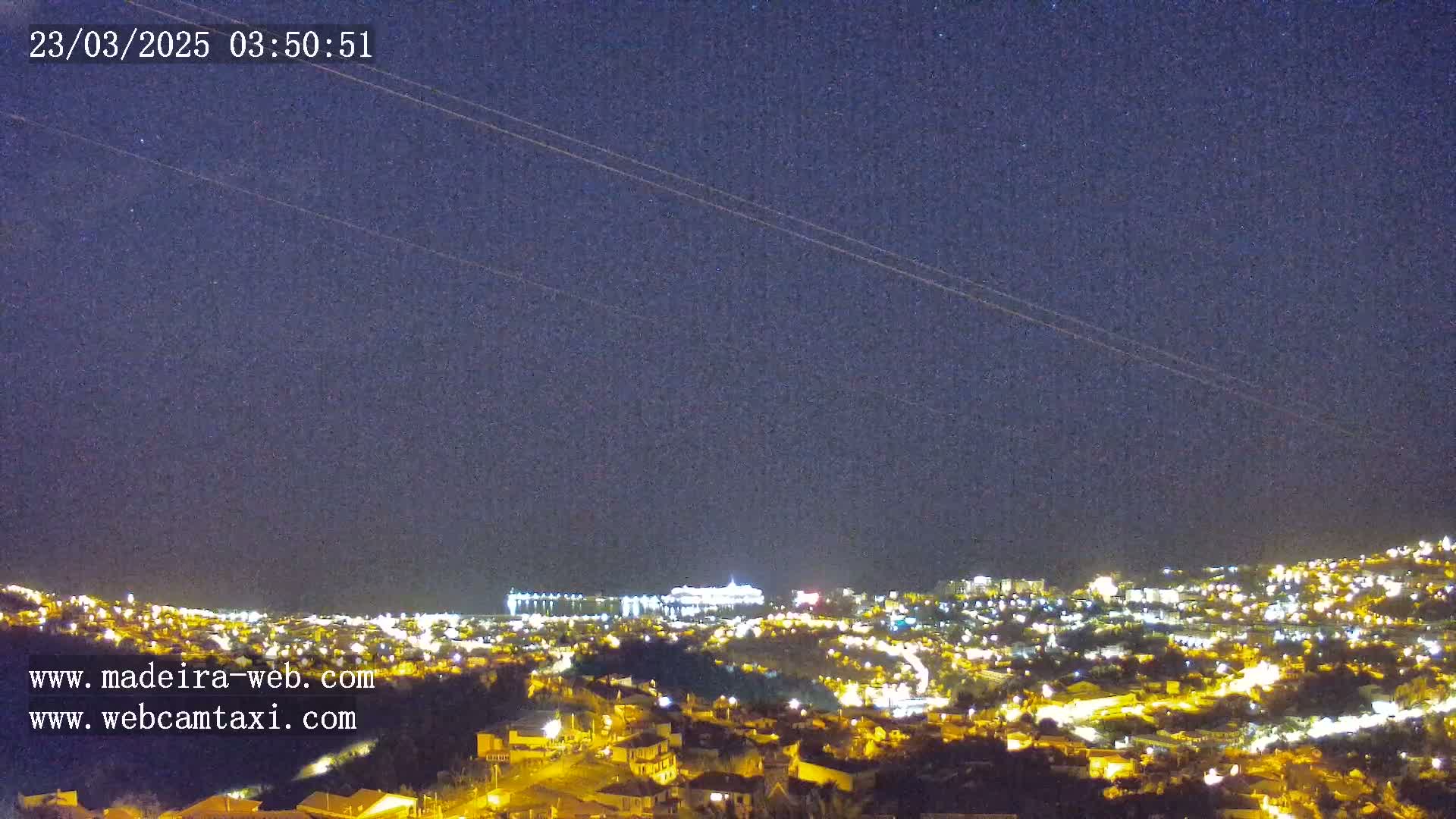 A nighttime long shot shows a city illuminated with many small lights under a dark, clear sky with a few stars visible and two airplane contrails.