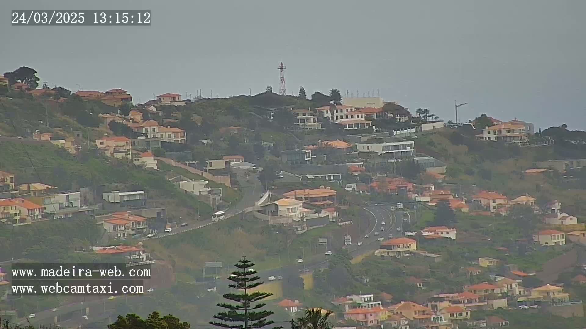 A hillside covered in houses and vegetation is visible under an overcast sky, with a highway curving through the scene.