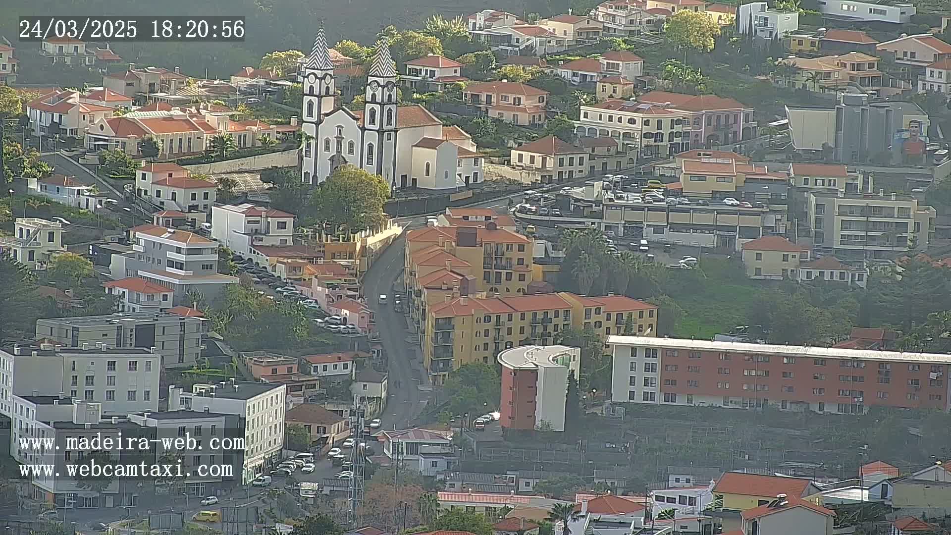 An aerial view shows a town with red-tiled roofs, a prominent white church with two towers, and buildings clustered on a hillside, under hazy conditions.