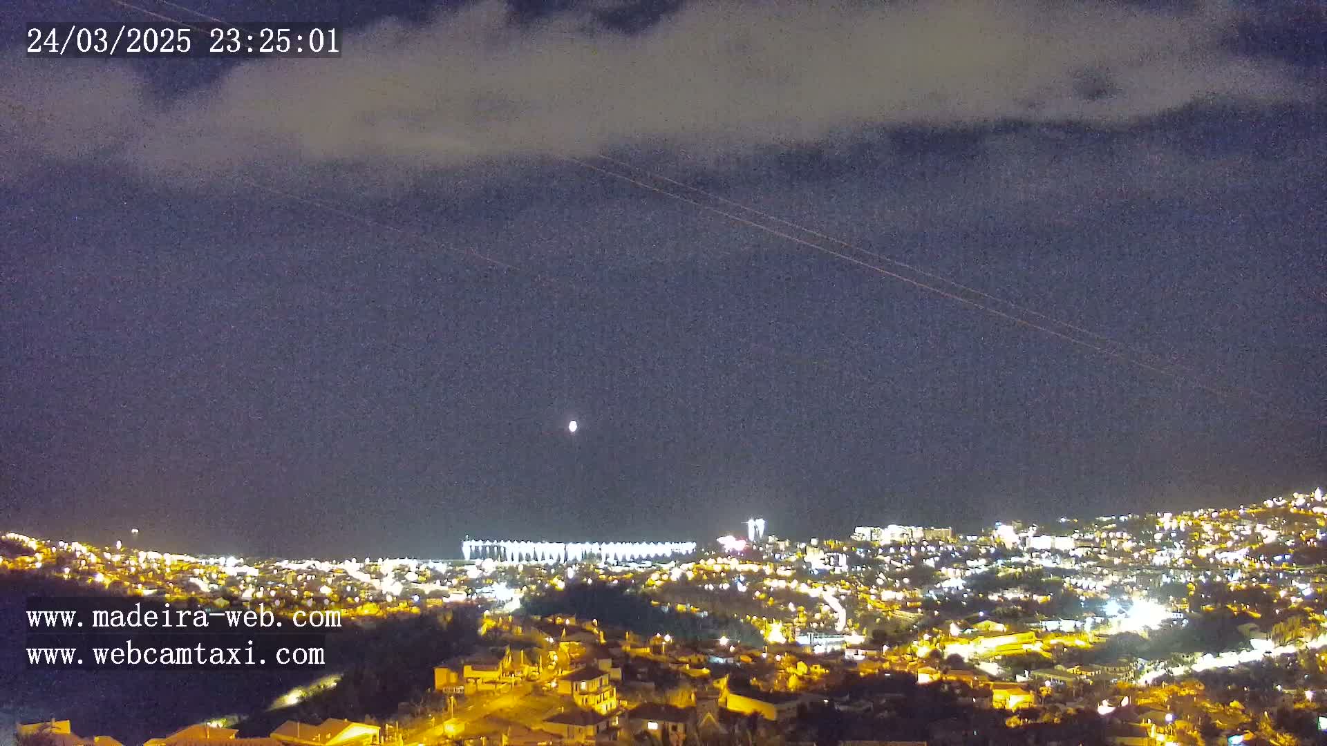 A nighttime view of a city illuminated by lights under a partly cloudy sky, with faint airplane trails visible.
