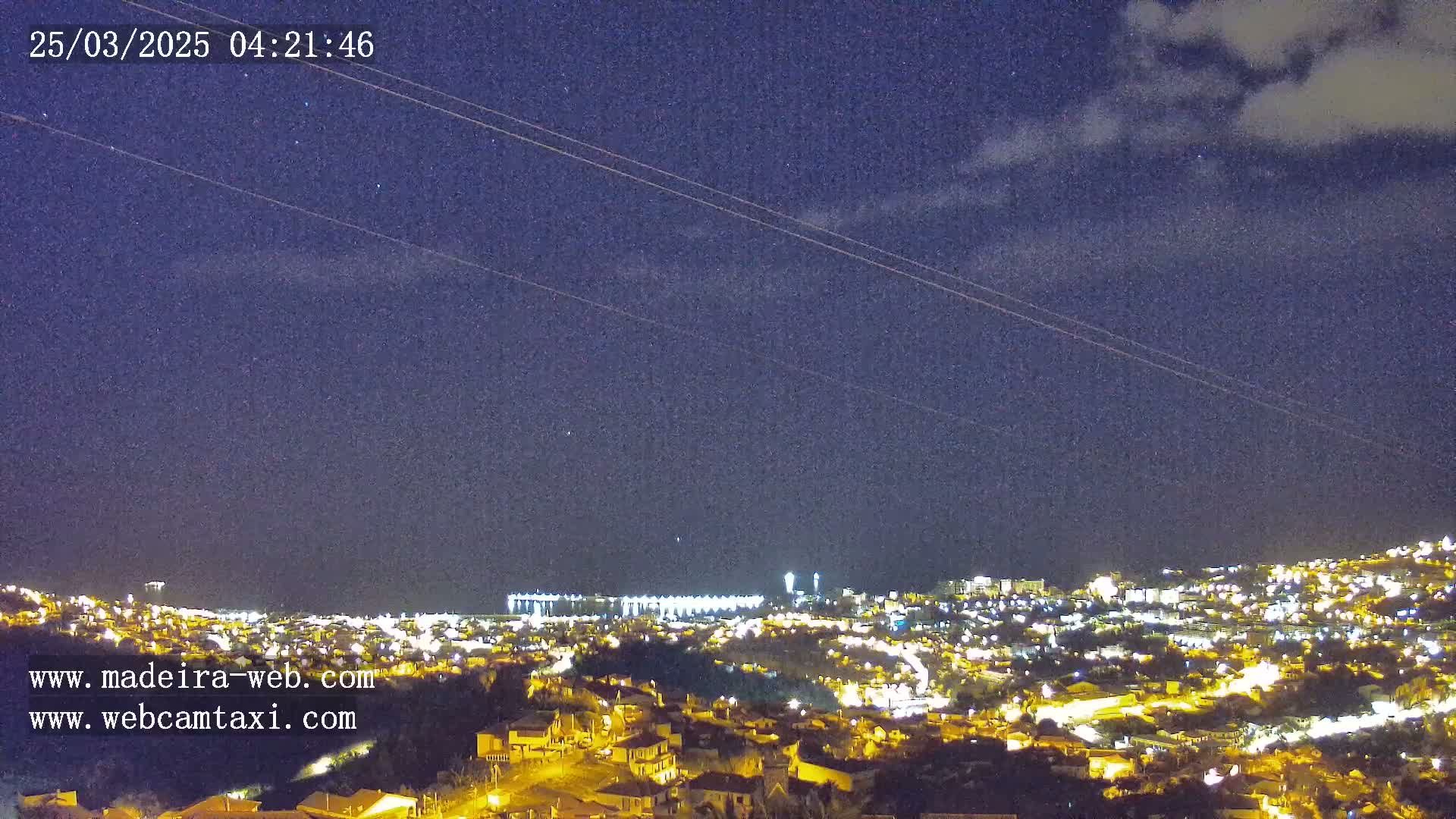 A nighttime view of a city illuminated by lights under a mostly clear, dark blue sky with a few clouds and faint streaks across it.