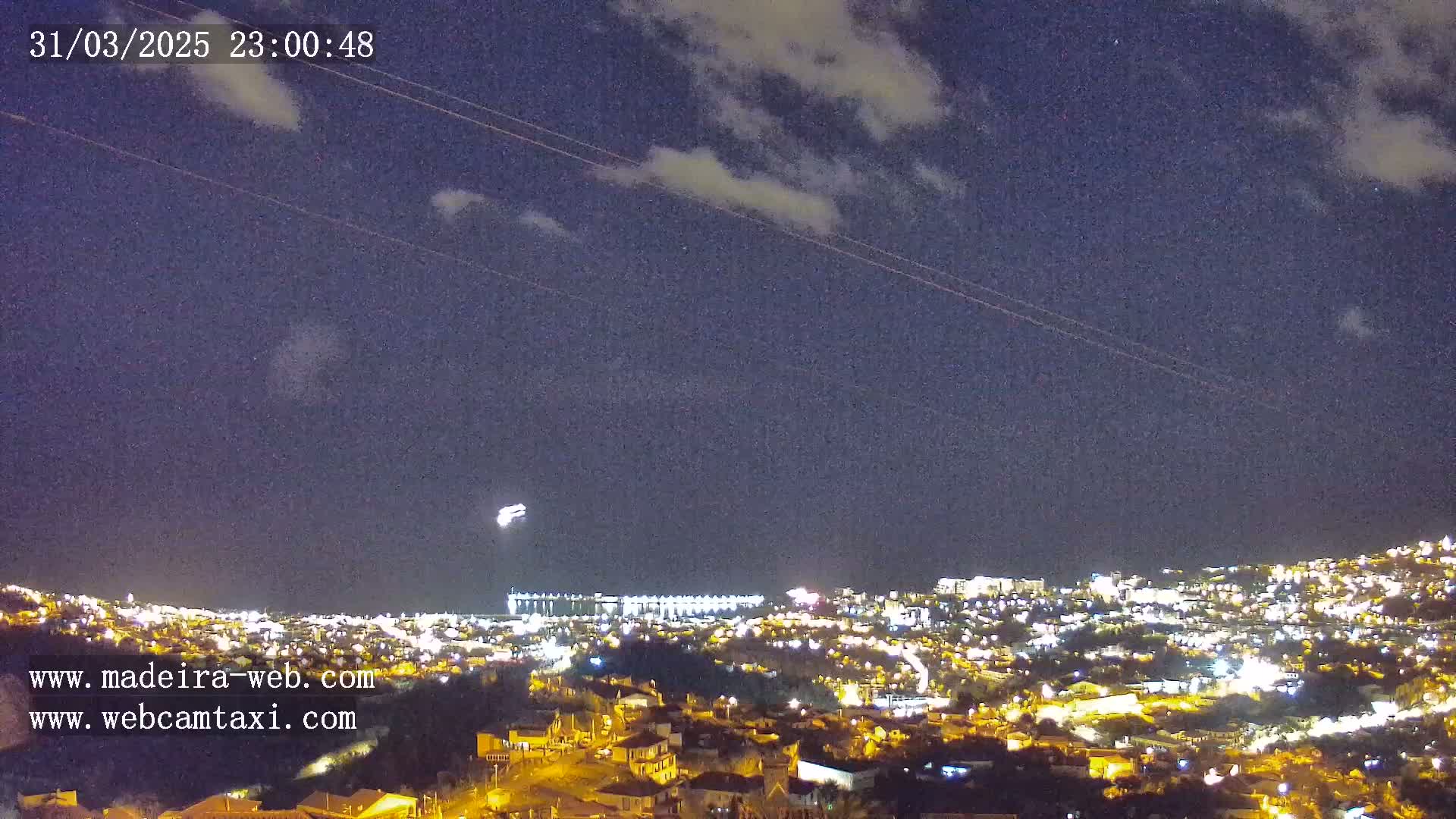 A nighttime view of a city illuminated by lights under a partly cloudy sky with faint contrails visible.