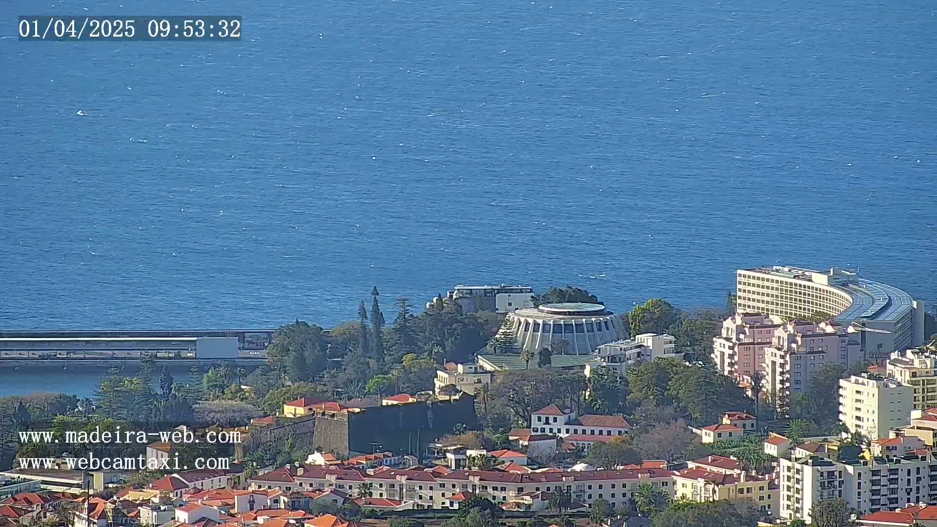 A coastal city with buildings of varying heights, including a large curved hotel, overlooks a calm blue ocean under a sunny sky.