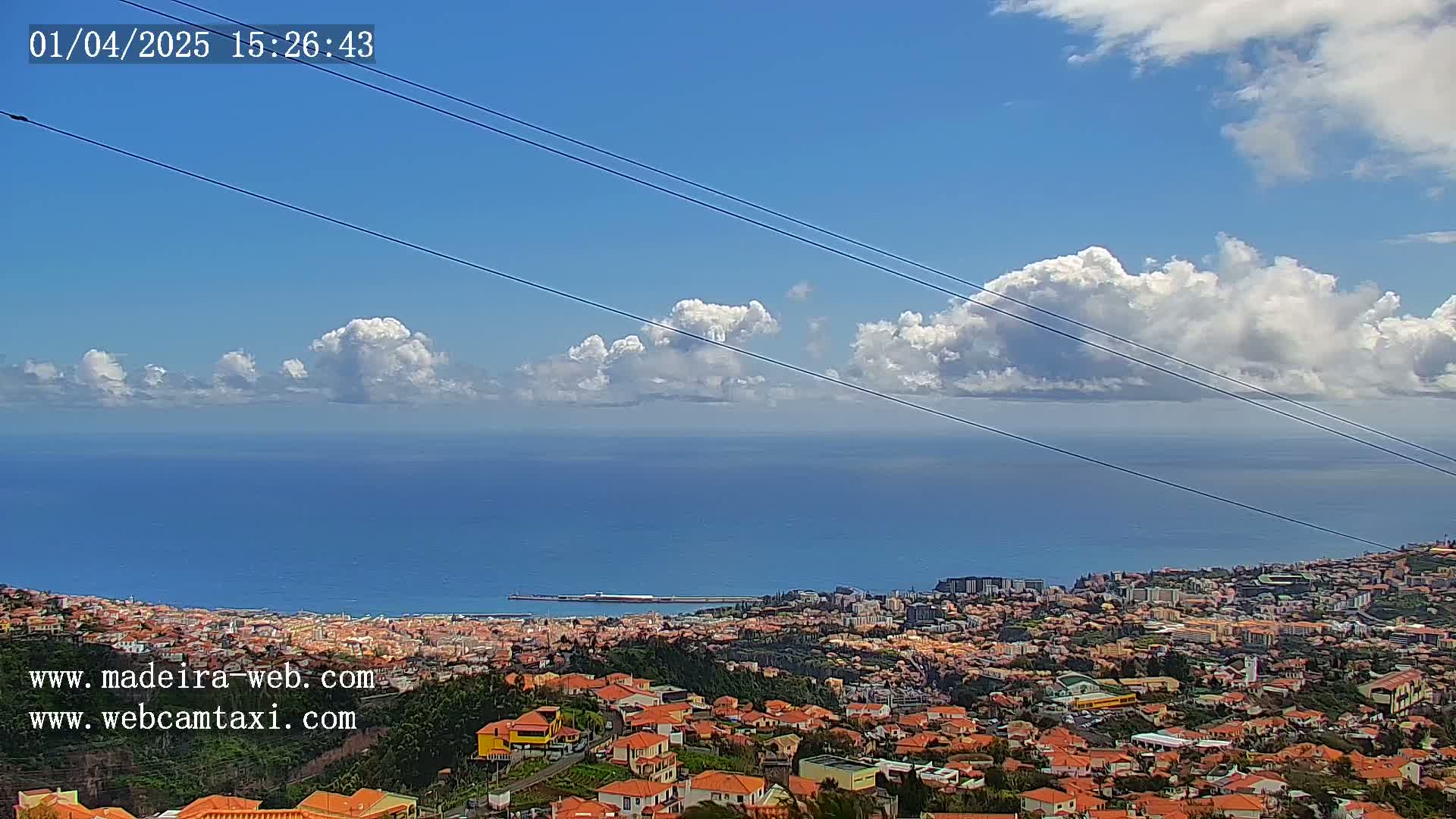 A coastal city with predominantly orange-roofed buildings stretches towards a calm ocean under a partly cloudy blue sky.