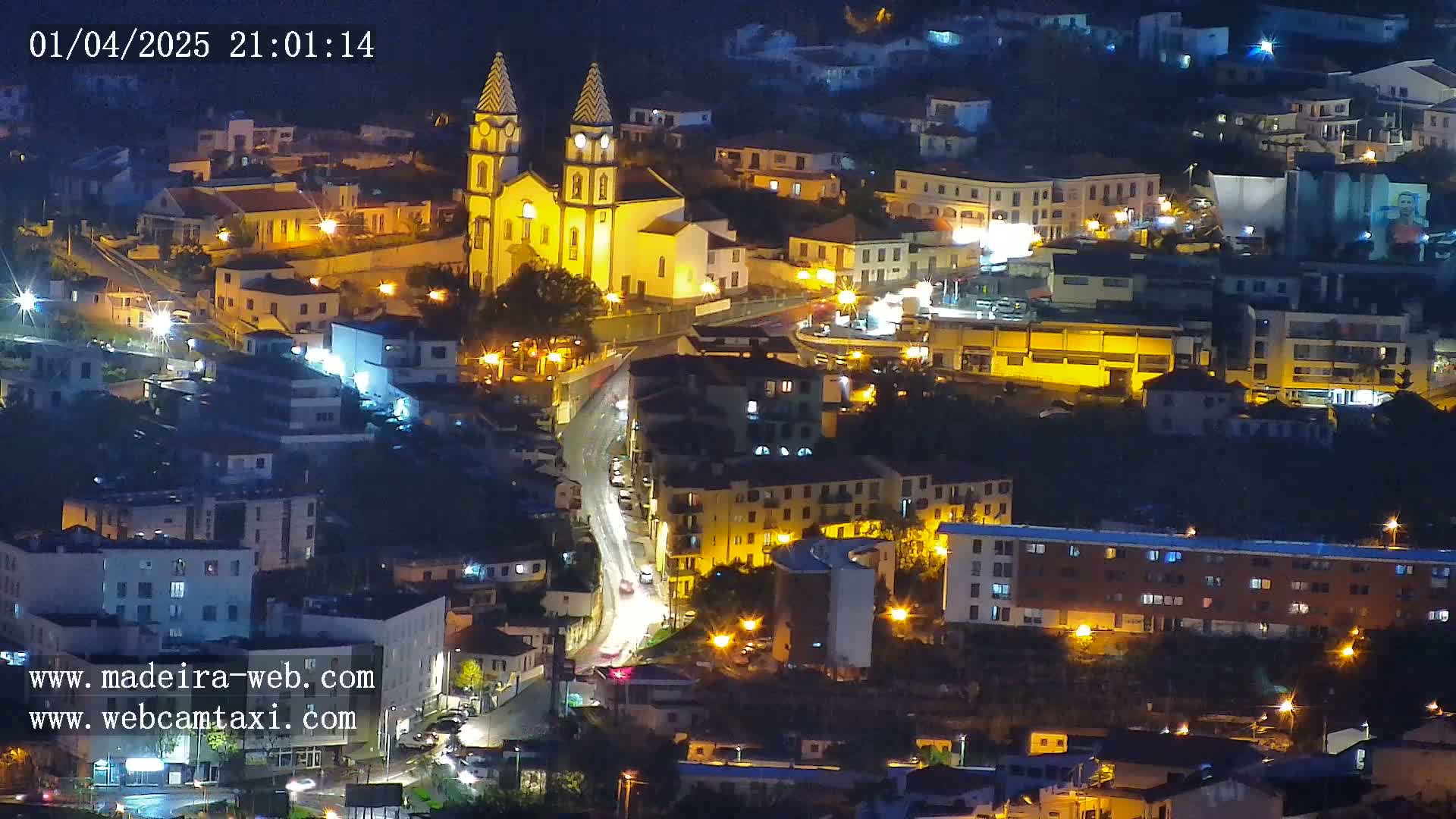 A nighttime aerial view of a town, illuminated by streetlights, features a prominent church at its center, with cars traveling along a winding road.