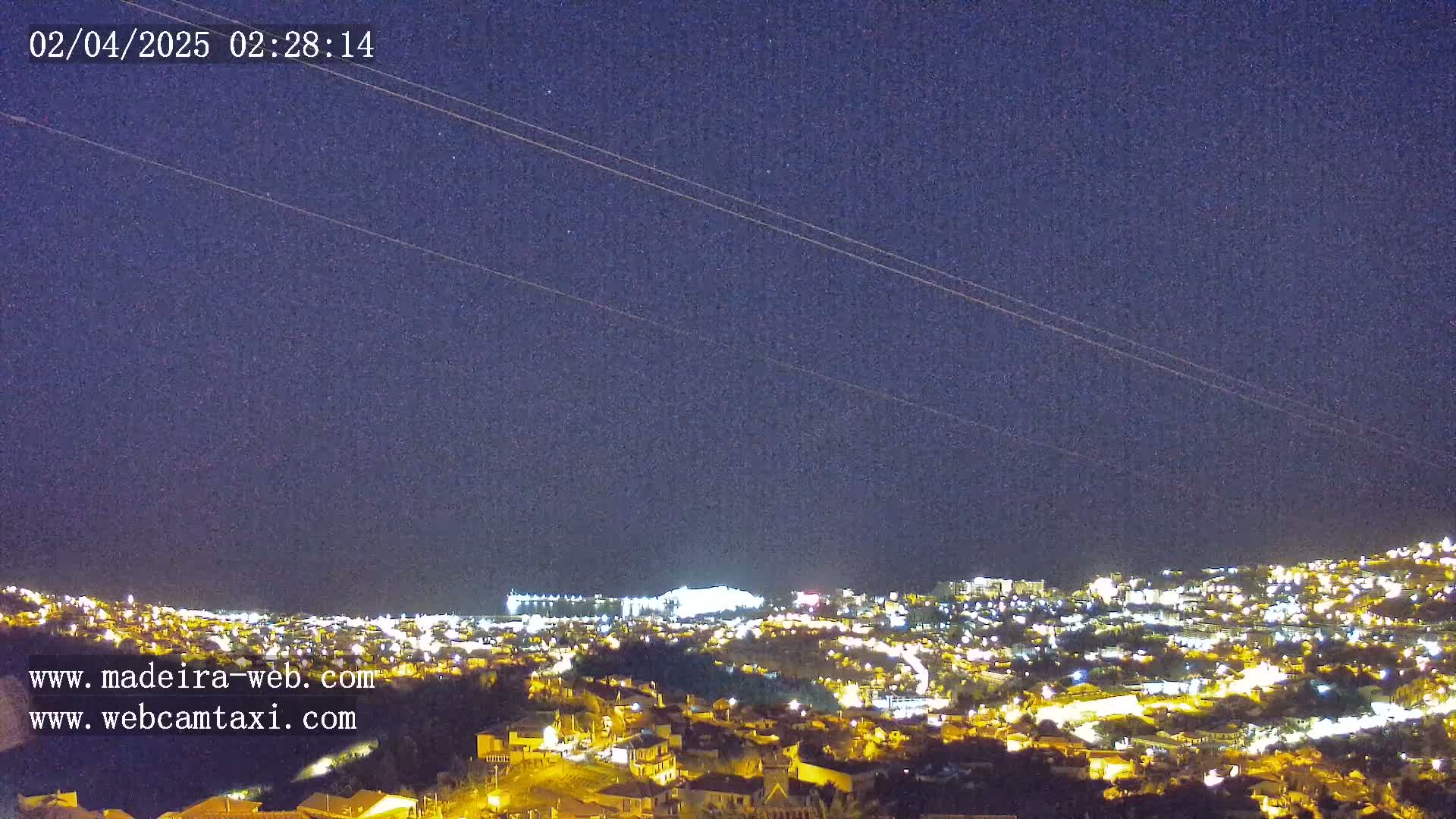 A nighttime, clear-sky view of a hillside city illuminated with lights, extending to a coastline in the distance.