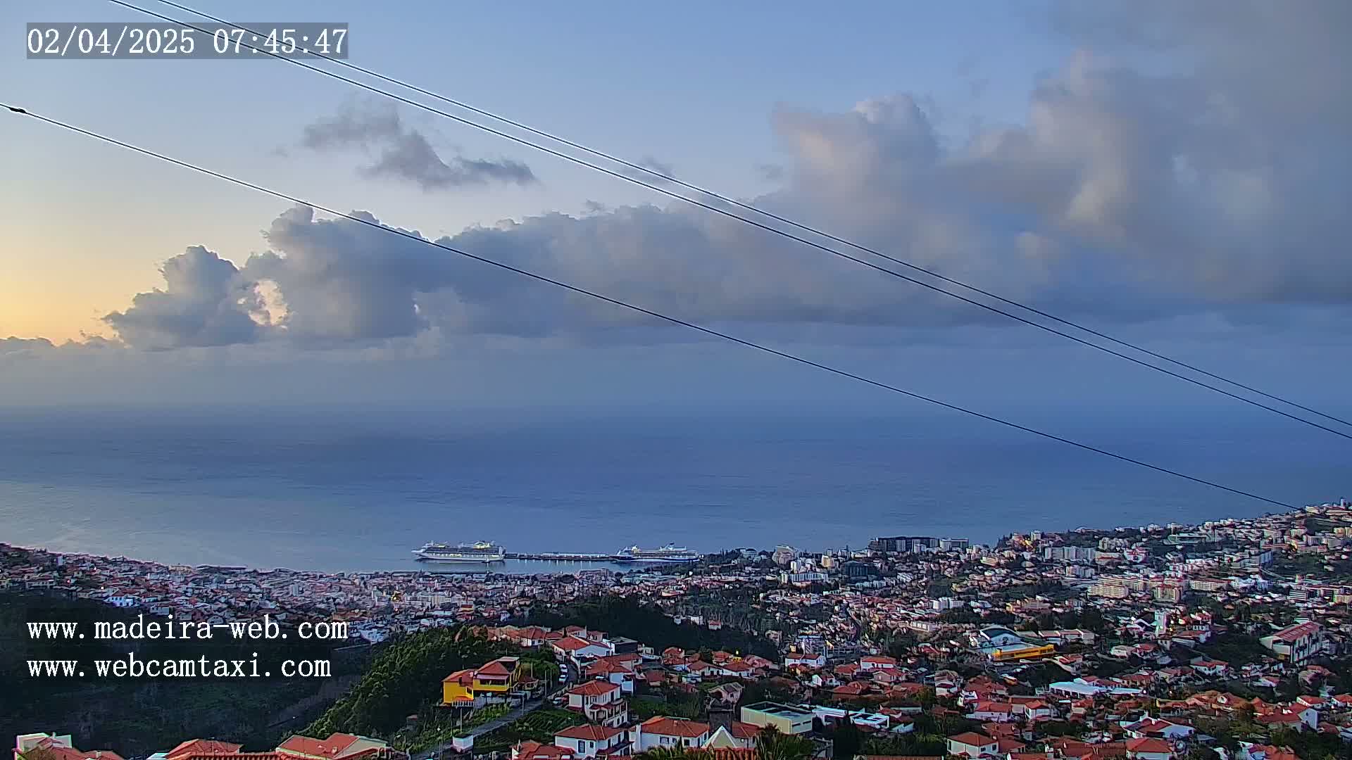 A coastal town with red-roofed houses sits below a partly cloudy sky, overlooking a calm ocean where a cruise ship is docked.