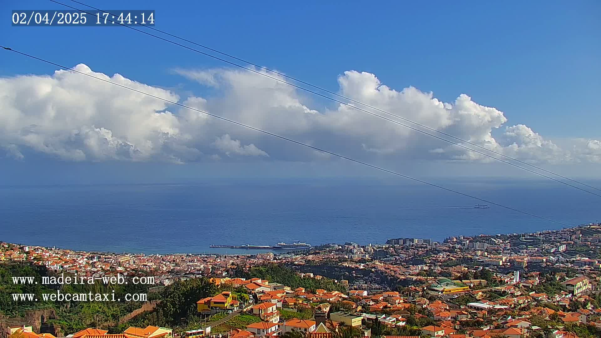 A coastal city with orange-roofed houses is nestled beneath a partly cloudy sky, overlooking a calm ocean with a large vessel in the distance.