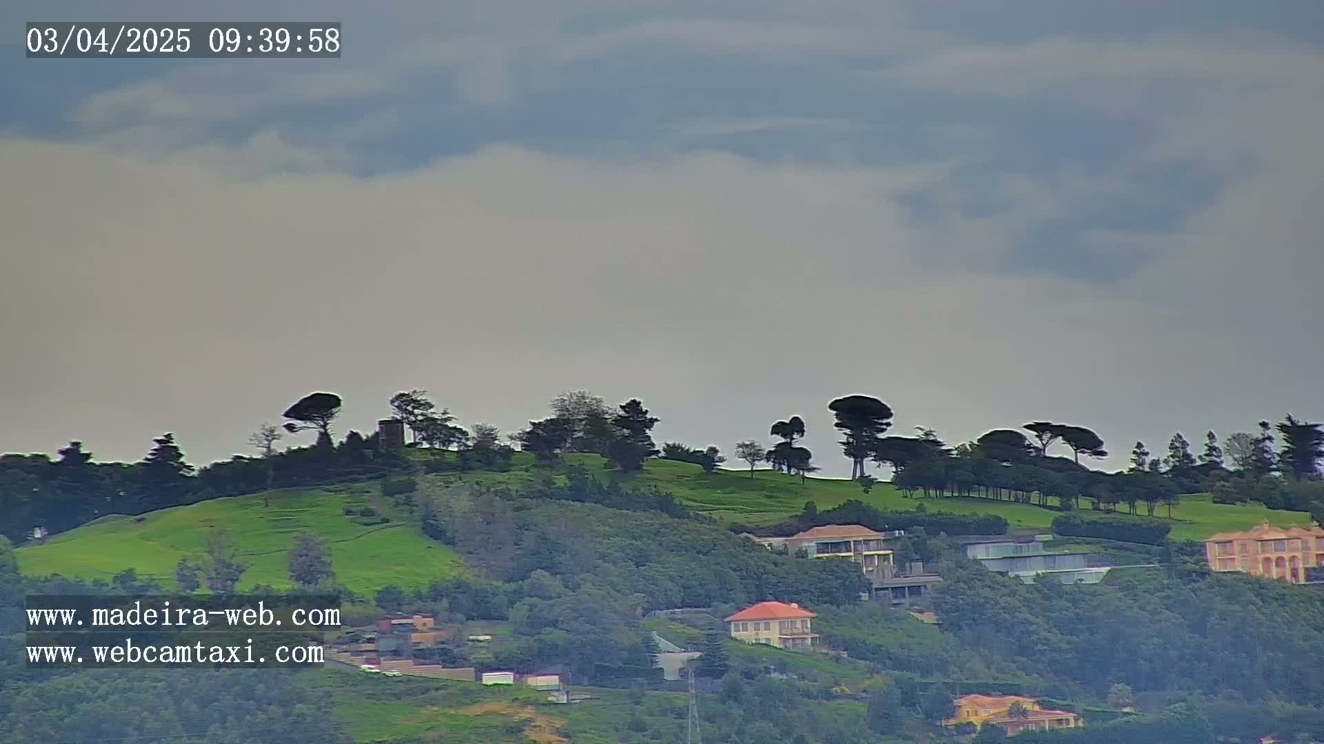 A mostly cloudy sky overlooks a verdant hillside dotted with trees and houses.