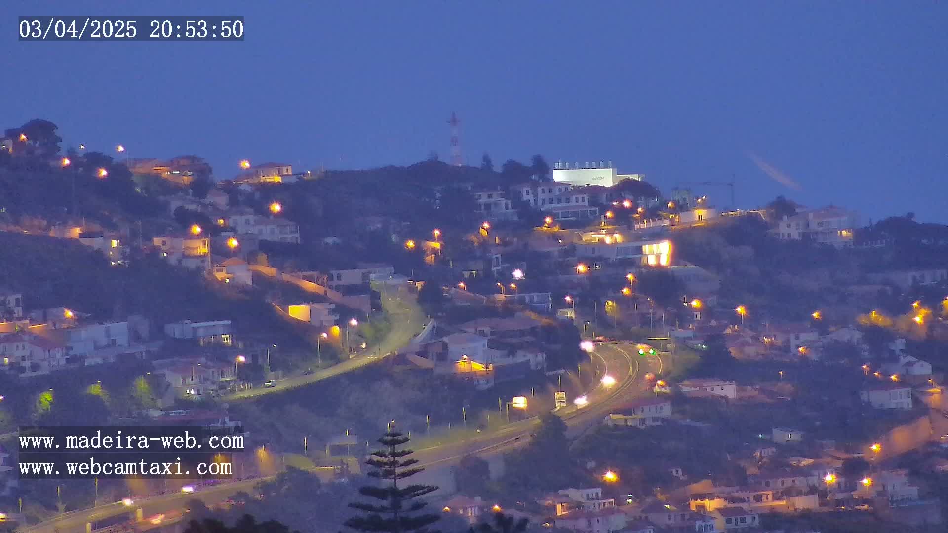 A nighttime view of a hillside town with illuminated houses and roads, under a dark blue, twilight sky.