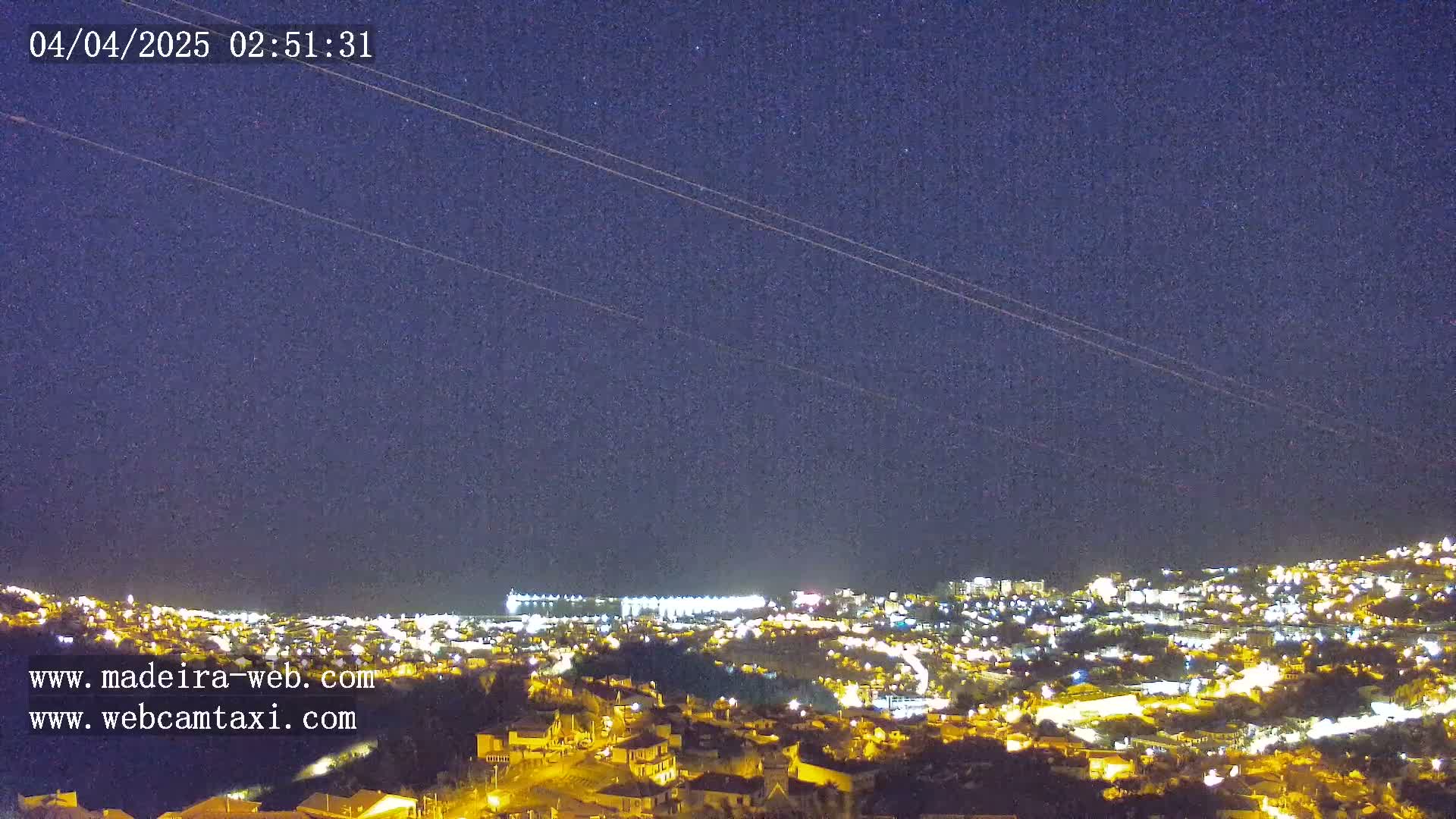 A nighttime view of a city illuminated by lights under a clear, dark sky with two faint trails across it.