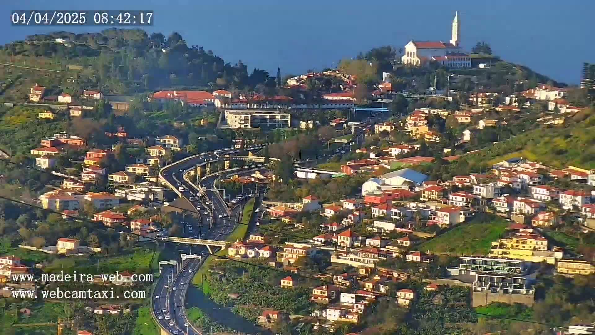 A coastal town with red-roofed houses sprawls across a hillside, a highway winds through it, and a church sits atop a nearby hill under a clear, sunny sky.
