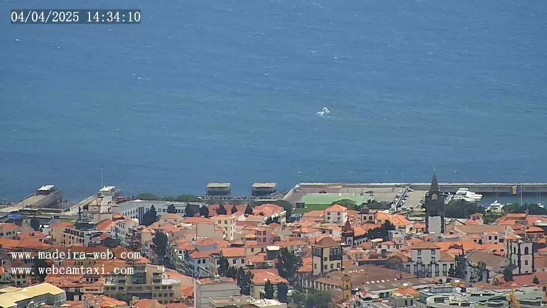 A coastal town with red-tiled roofs and a prominent church tower is situated next to a calm blue ocean under a clear sky.