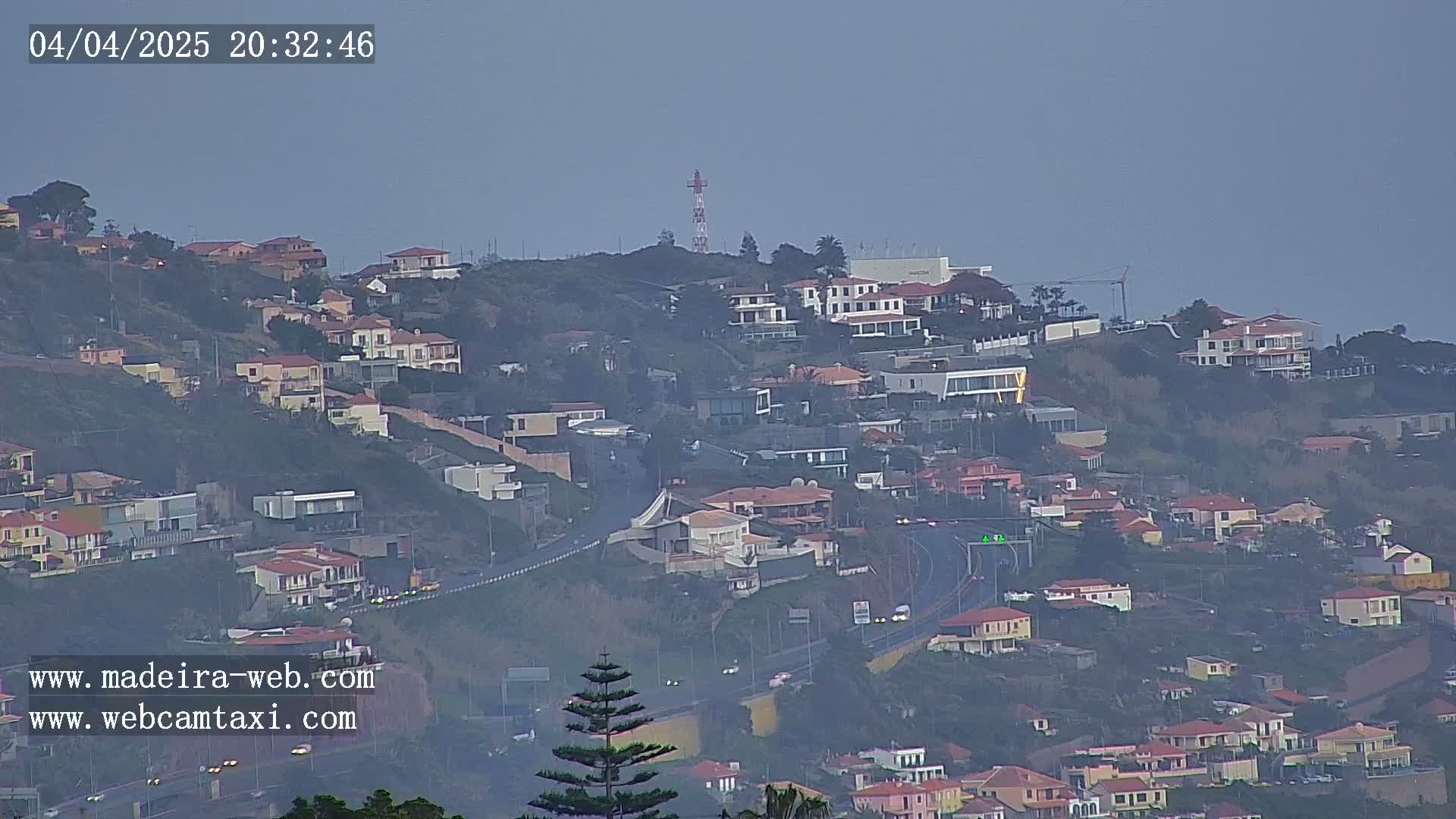 A hazy view of a hillside residential area with a winding road, featuring numerous houses and a communications tower in the distance.