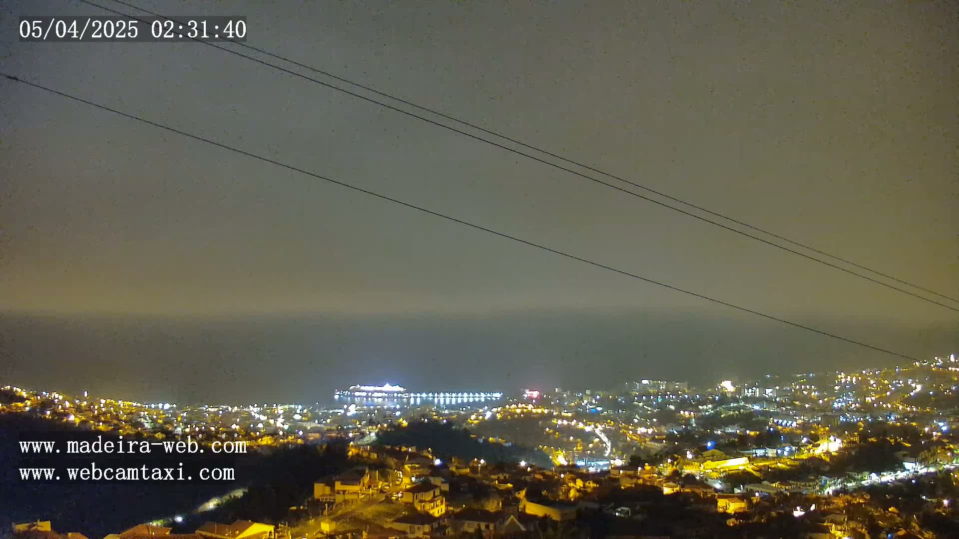A nighttime, hazy view overlooking a coastal city illuminated with lights, featuring a large ship at the harbor.