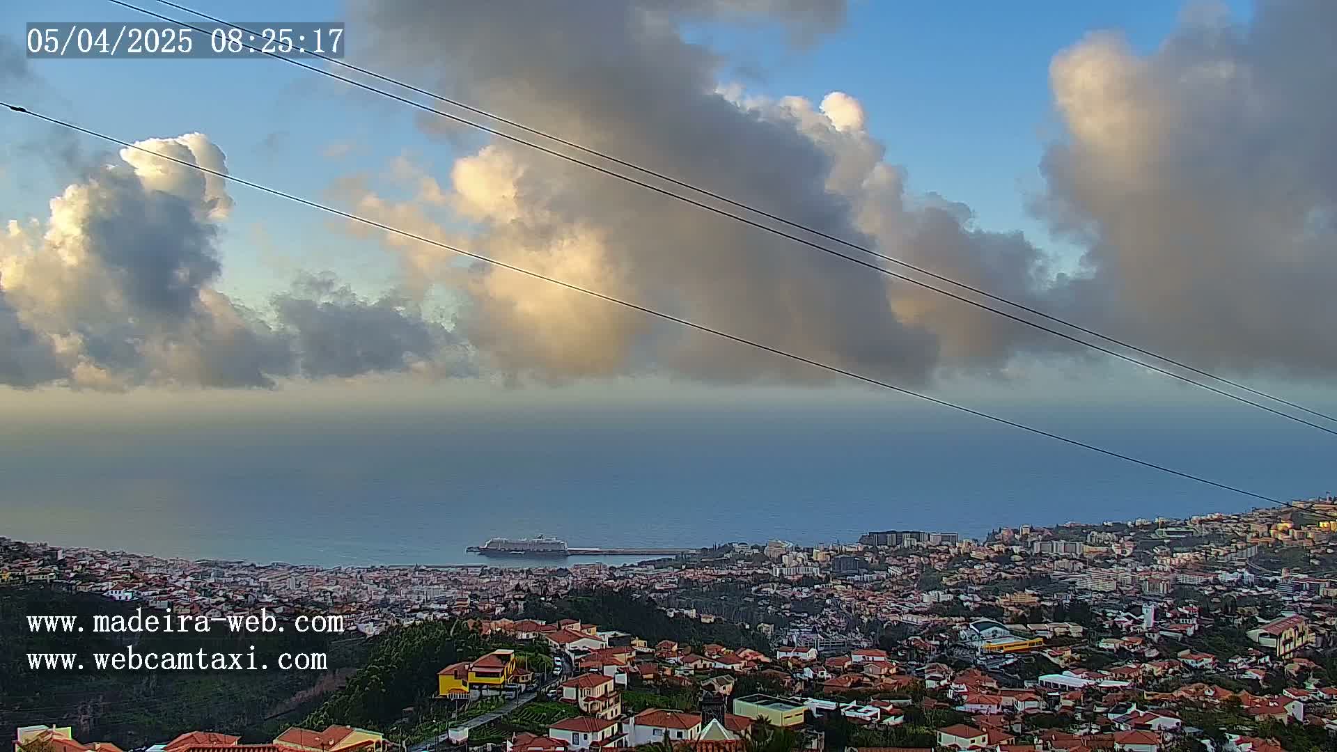 A coastal city with red-roofed houses sits below a partly cloudy sky, overlooking a calm ocean with a cruise ship in the distance.