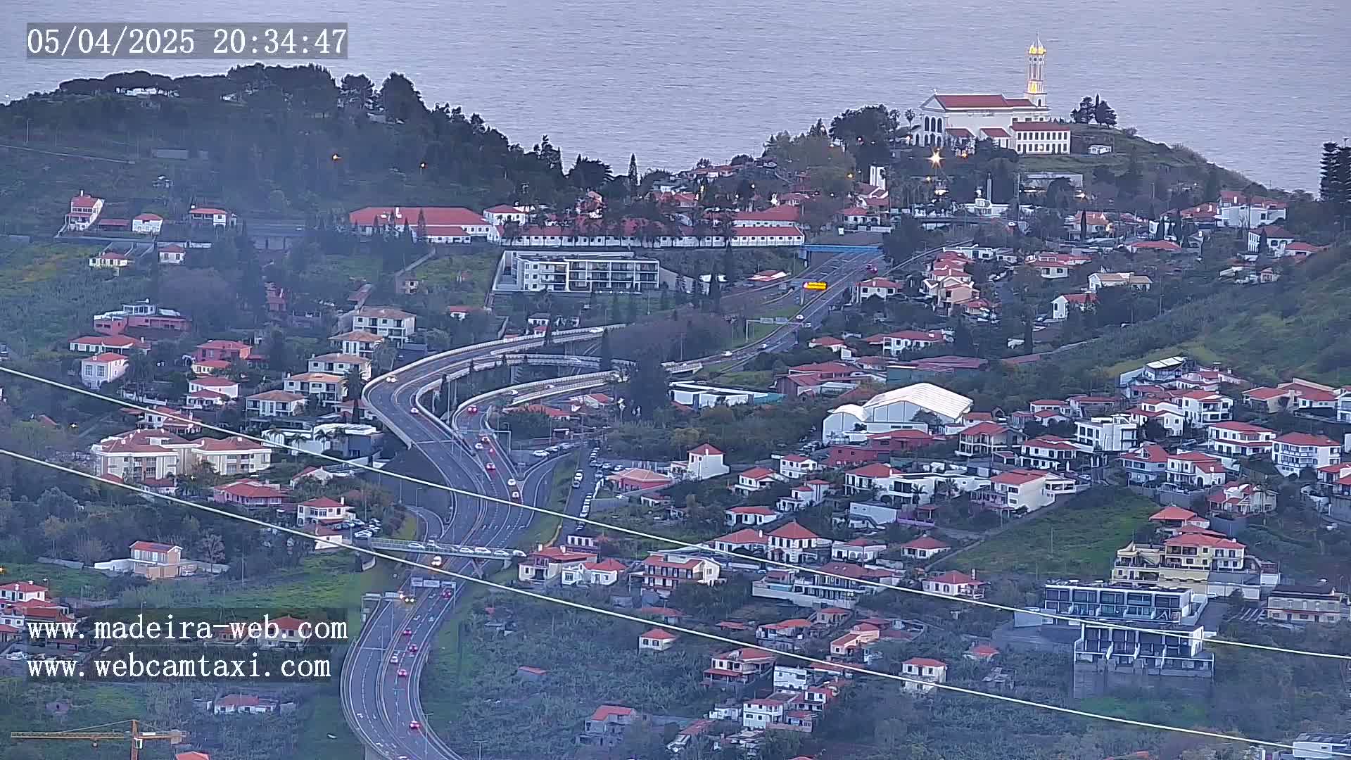 An elevated view shows a coastal town with red-roofed houses clustered on a hillside, overlooking a highway and a calm ocean under a dusky sky.