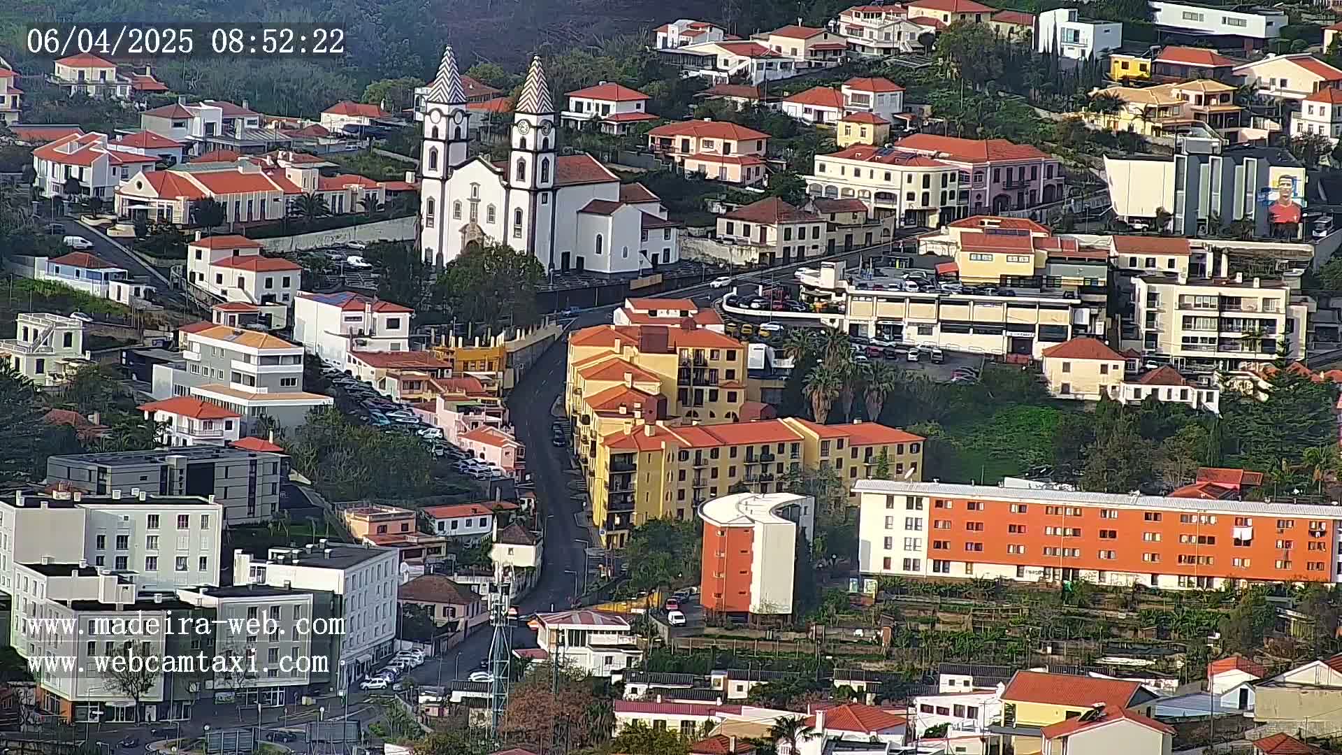 An aerial view shows a town with predominantly red-tiled roofs, a prominent white church with tall spires, and several multi-story apartment buildings, all situated on a hillside under a clear sky.