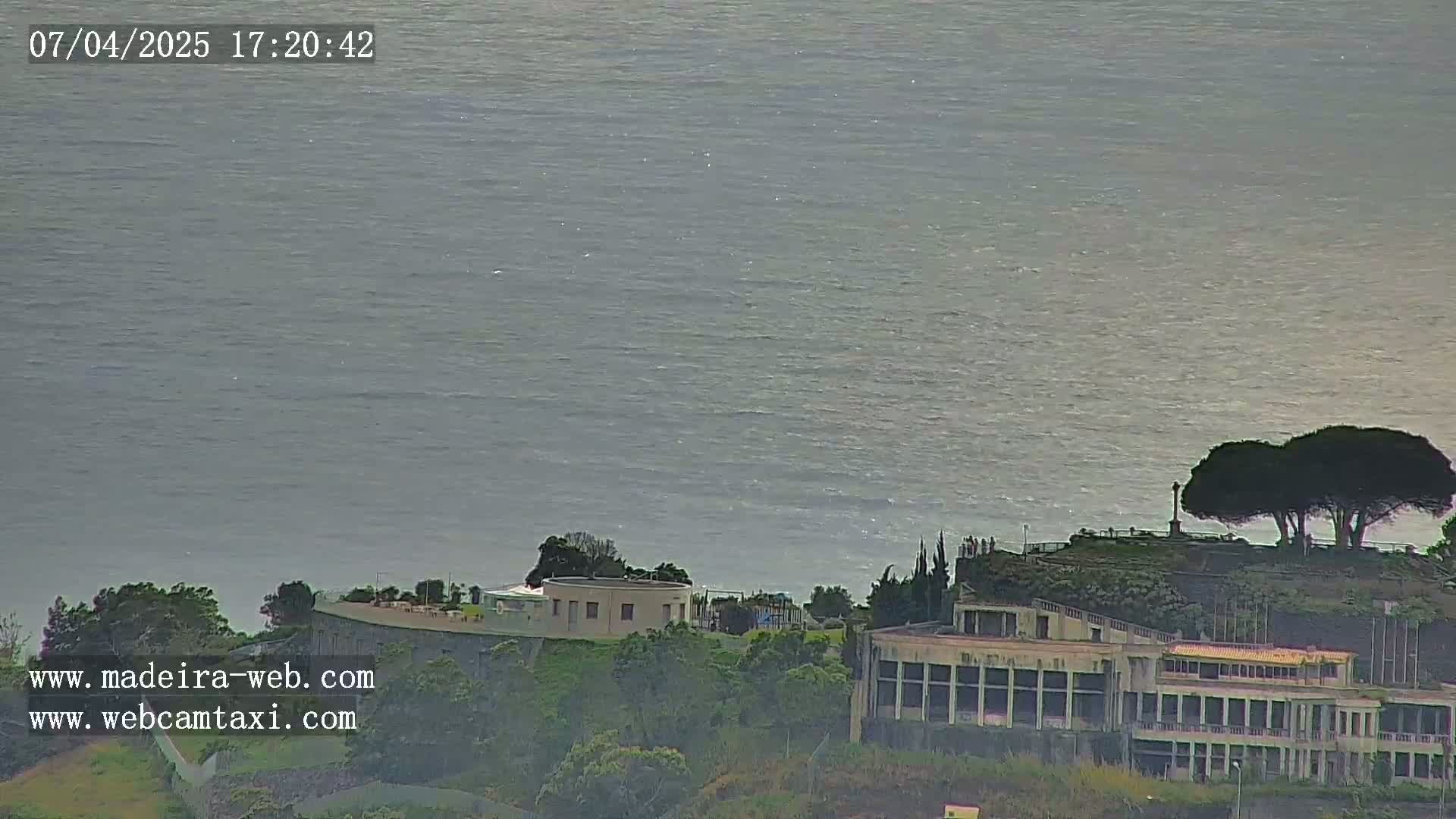 A calm, gray ocean stretches to the horizon behind a coastal building complex and a few trees atop a small hill.