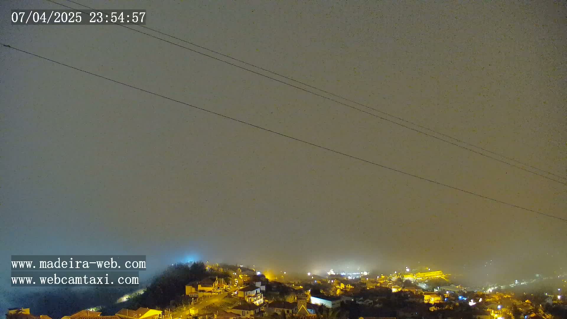 A nighttime, foggy view shows a hillside town with scattered lights under a dark sky, crisscrossed by power lines.