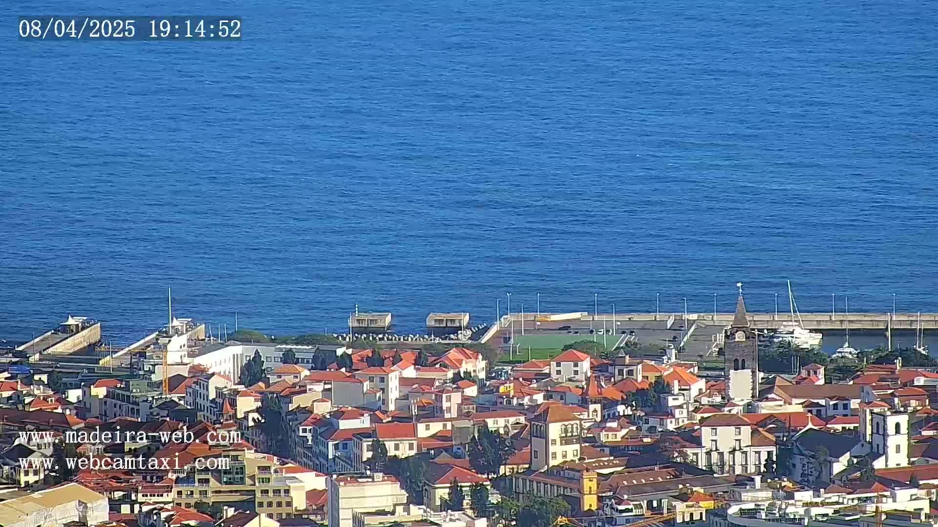 A coastal town with red-roofed buildings and a small harbor is visible on a sunny day, overlooking a calm blue ocean.