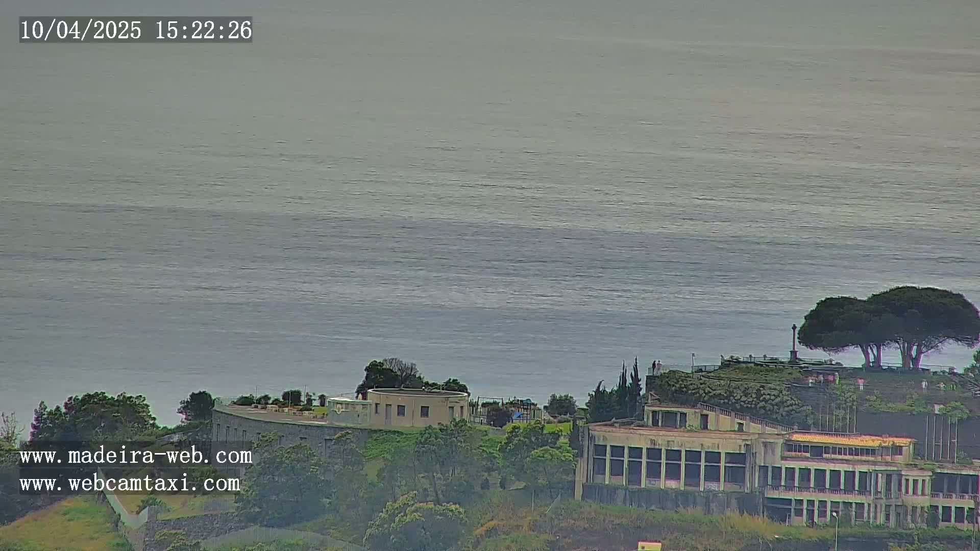 A large body of calm, gray-blue water is seen in the distance, beyond a coastal area featuring buildings, trees, and a small hilltop structure.
