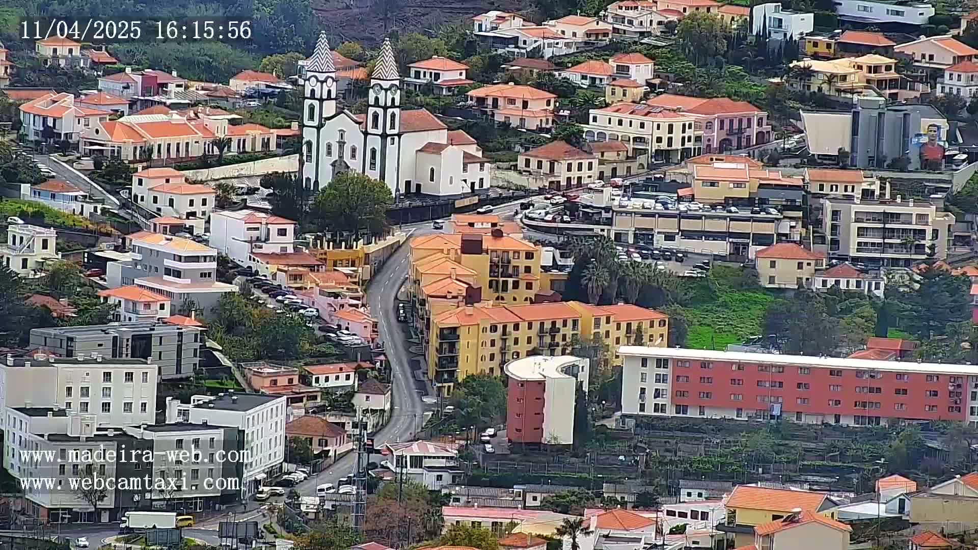 An aerial view shows a town with many low-rise buildings, a prominent church with twin towers, and a winding road, all under an overcast sky.