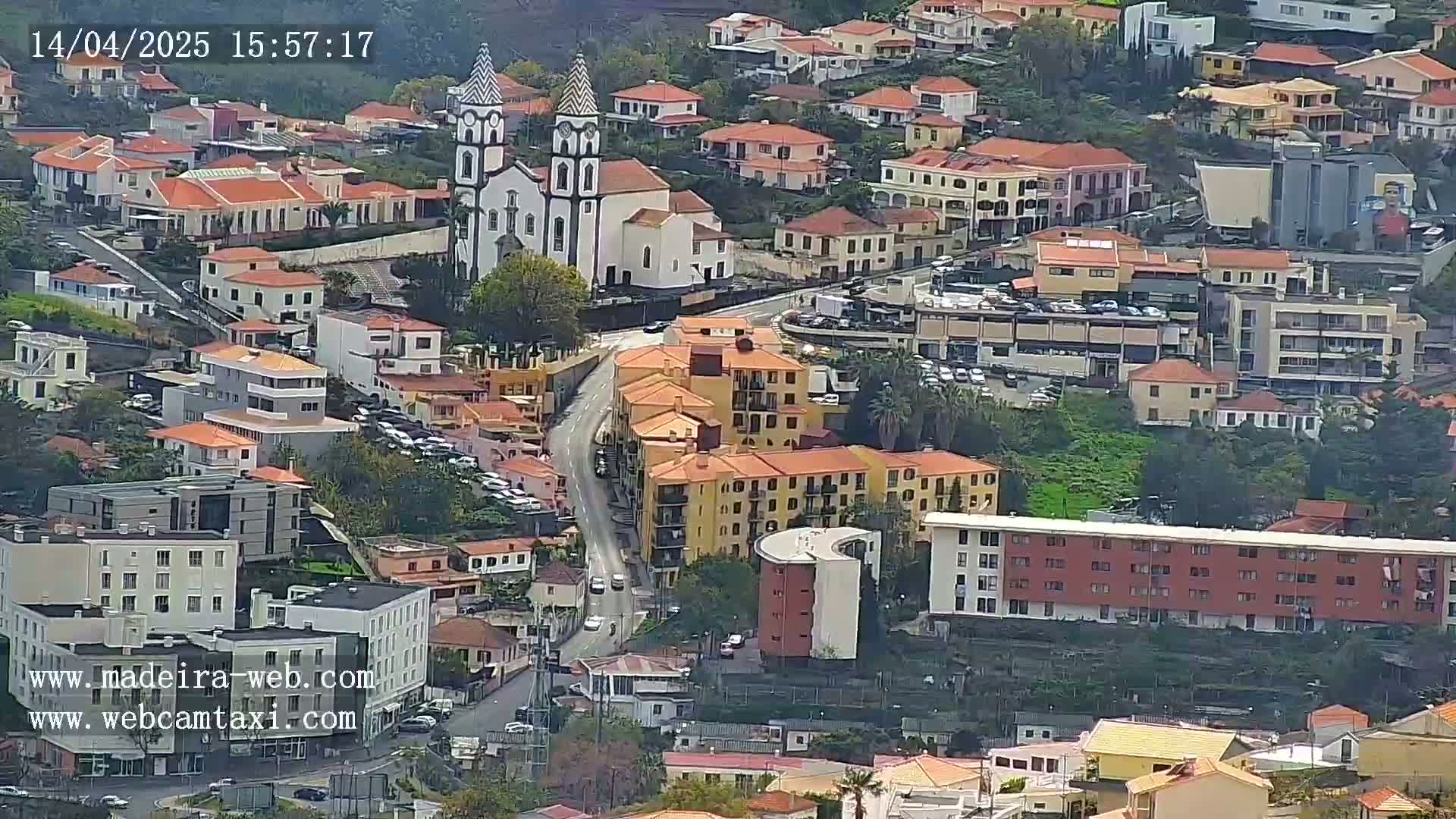 An aerial view shows a town with primarily low-rise buildings, a prominent church, and winding roads, under partly cloudy conditions.