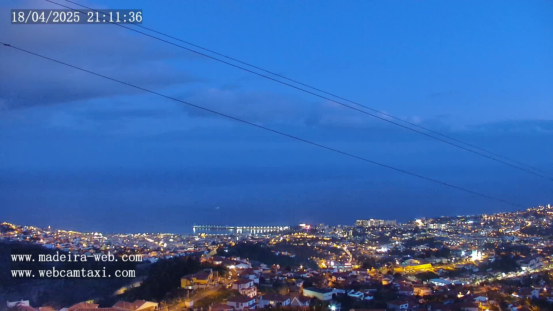 A nighttime view of a coastal city illuminated with lights under a mostly clear, dark blue twilight sky.
