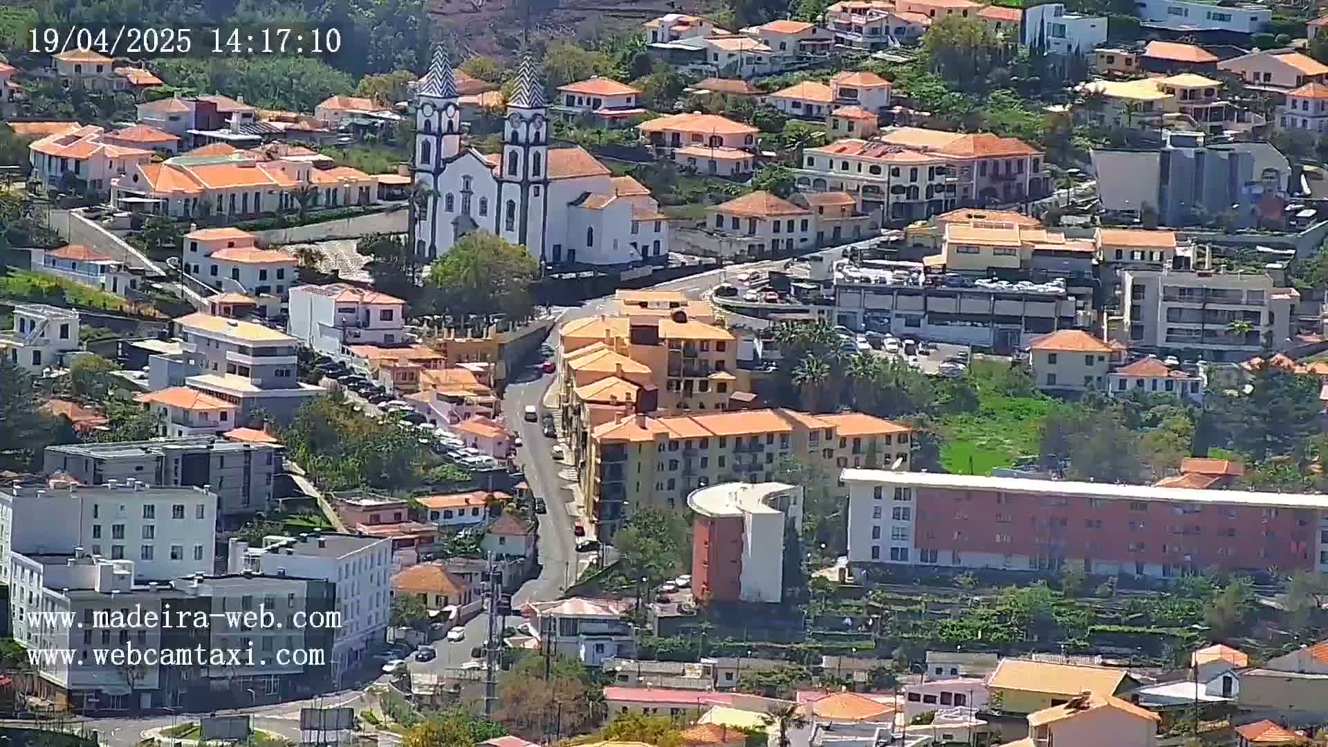 A sunny aerial view of a town shows numerous low-rise buildings with orange roofs clustered around a prominent white church with tall towers.