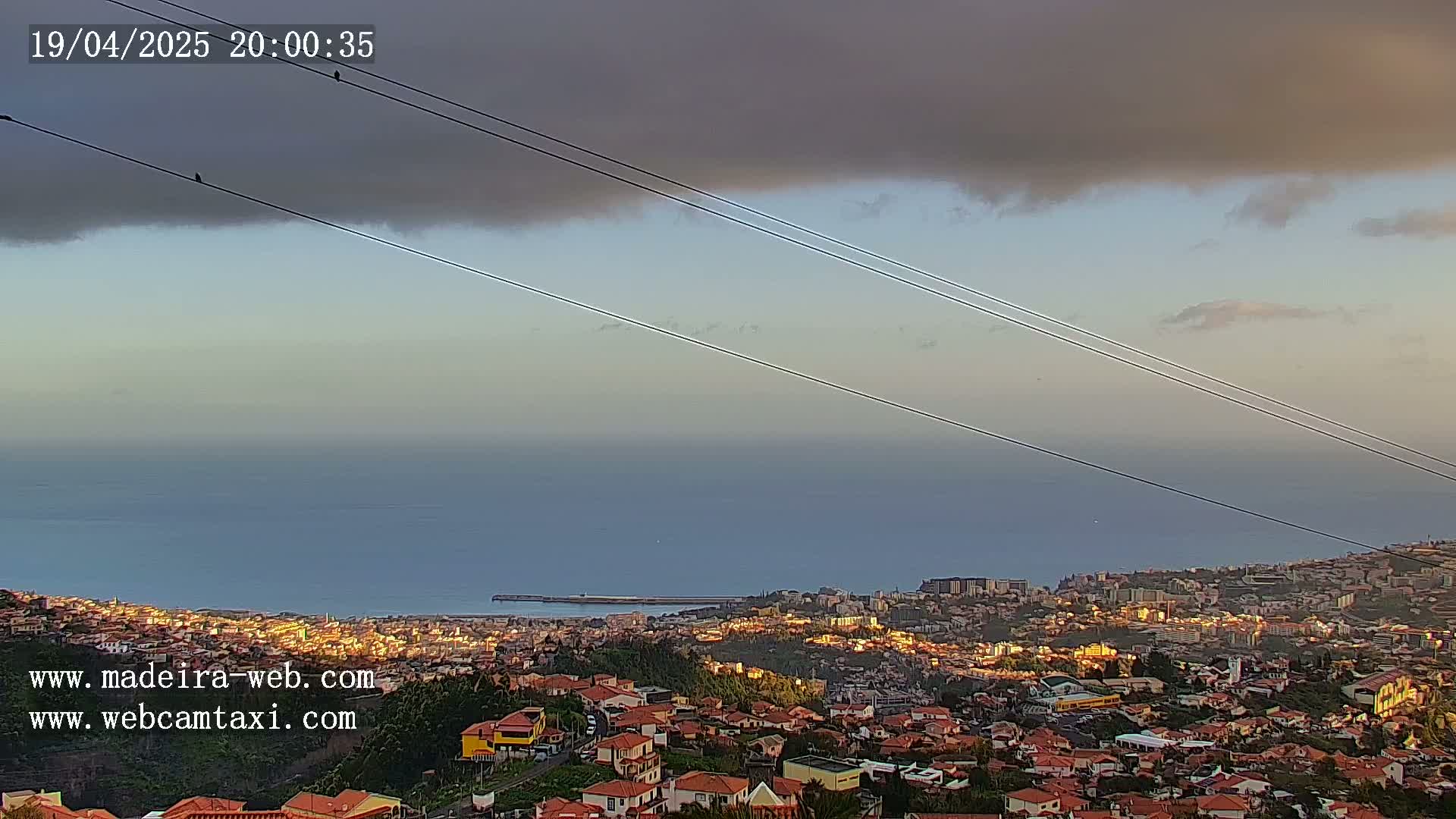 A coastal town with red-roofed houses extends to a calm ocean under a partly cloudy evening sky, with power lines stretching across the scene.