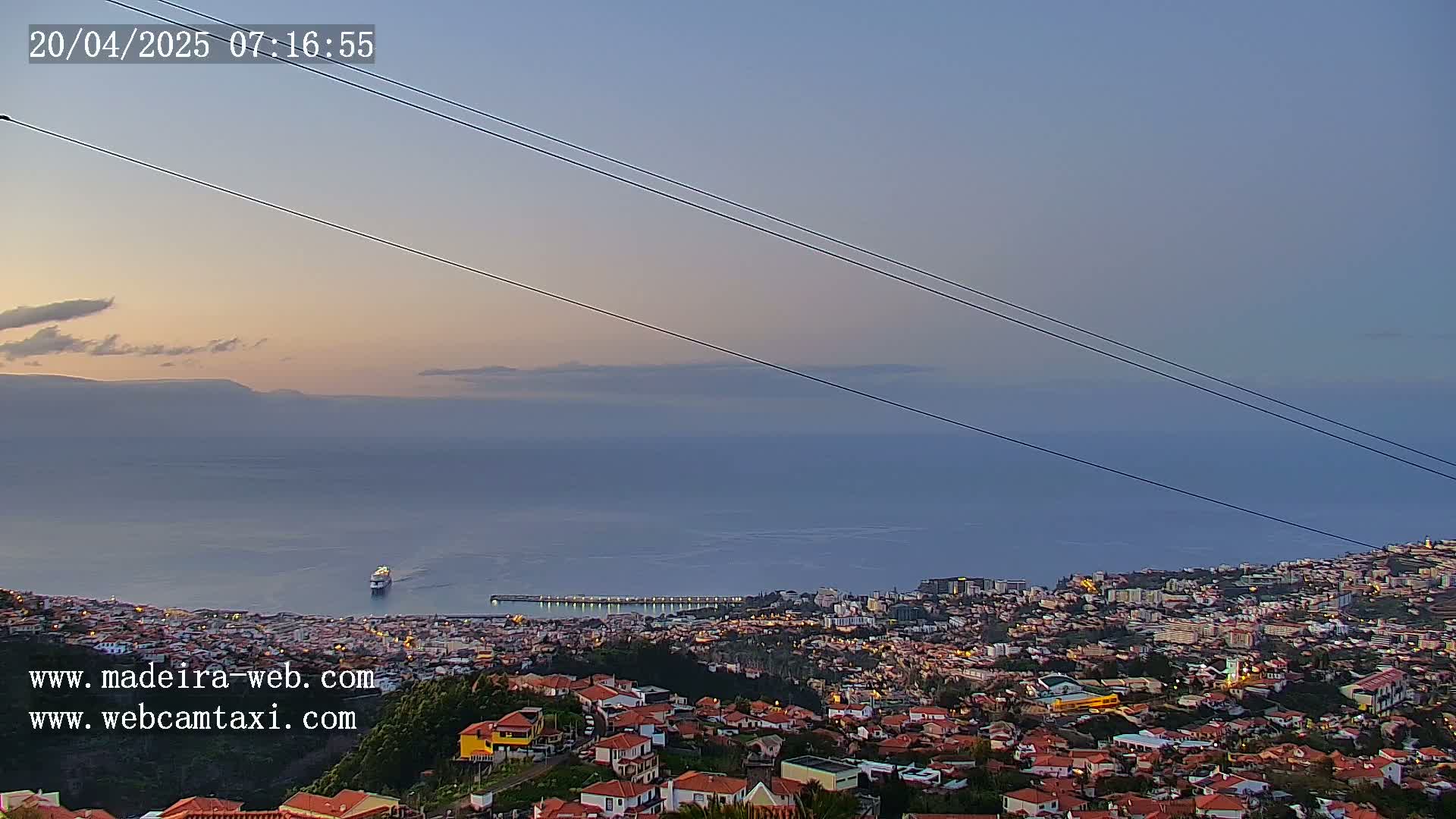 A coastal town at twilight is seen from a high vantage point, with a cruise ship visible in the calm ocean below.