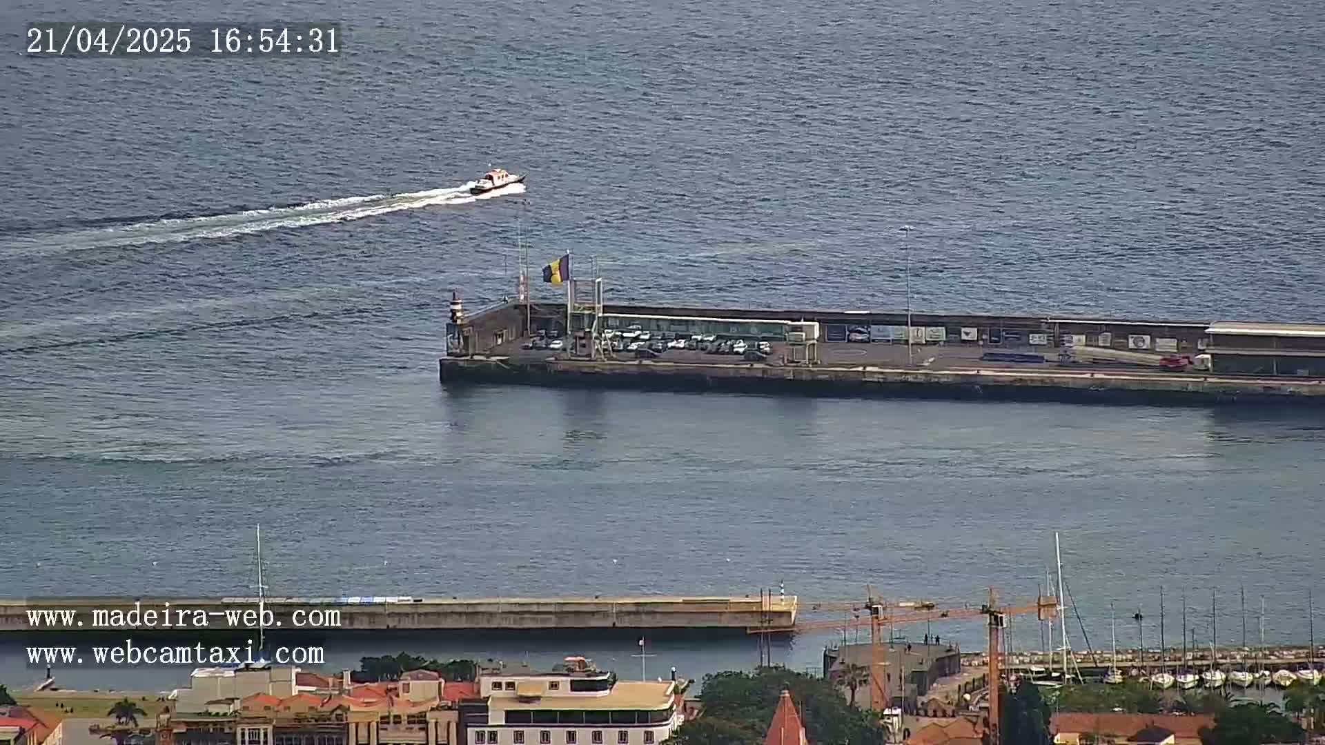 A small boat travels across a calm body of water toward a pier with a parking lot and a flag, next to a marina filled with boats.