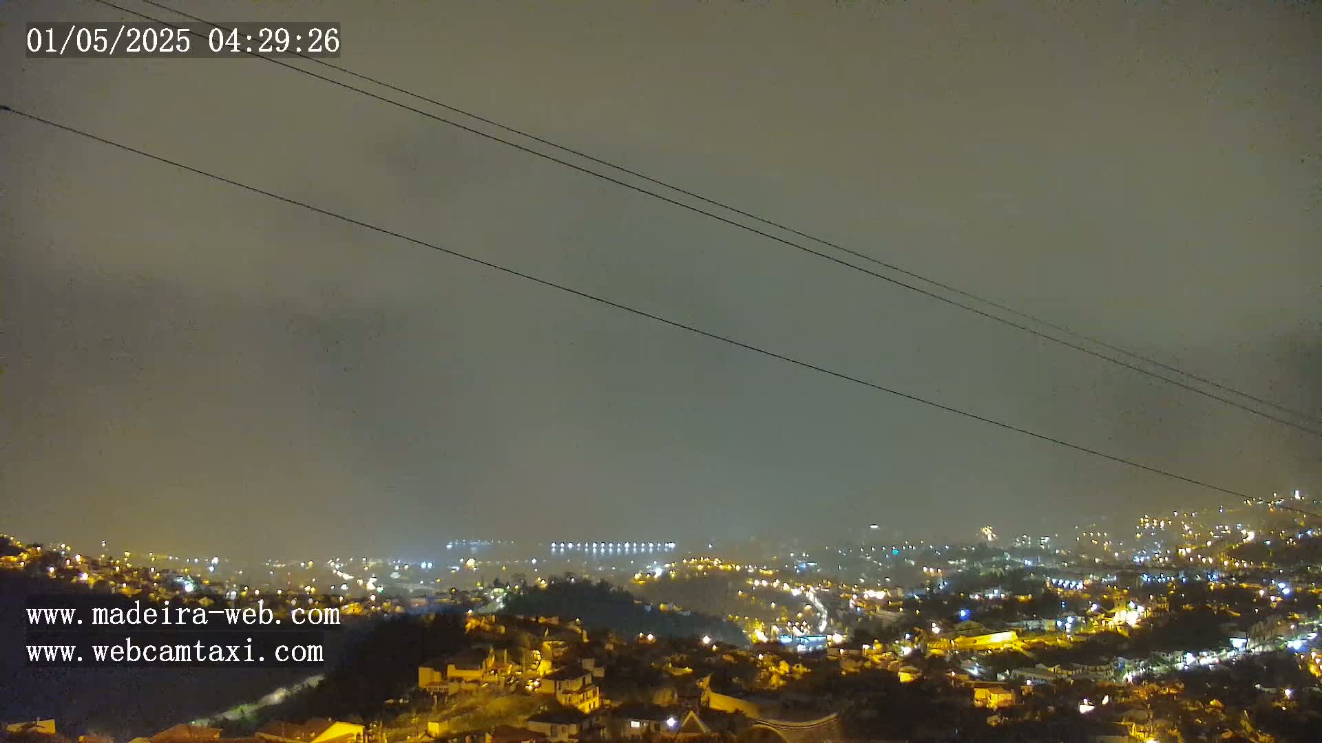 A hazy night scene shows a city illuminated with scattered lights below a dark, overcast sky, crisscrossed by power lines.