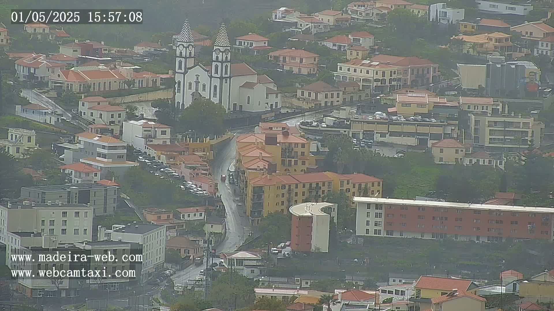 A rain-soaked town, viewed from above, features a prominent church with twin towers and numerous buildings clustered along curving streets.