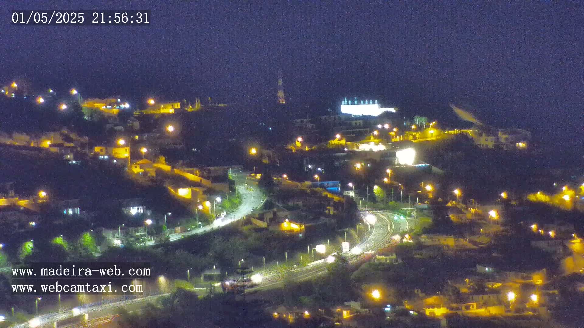 A nighttime view of a hilly town with illuminated roads and buildings under a dark sky.