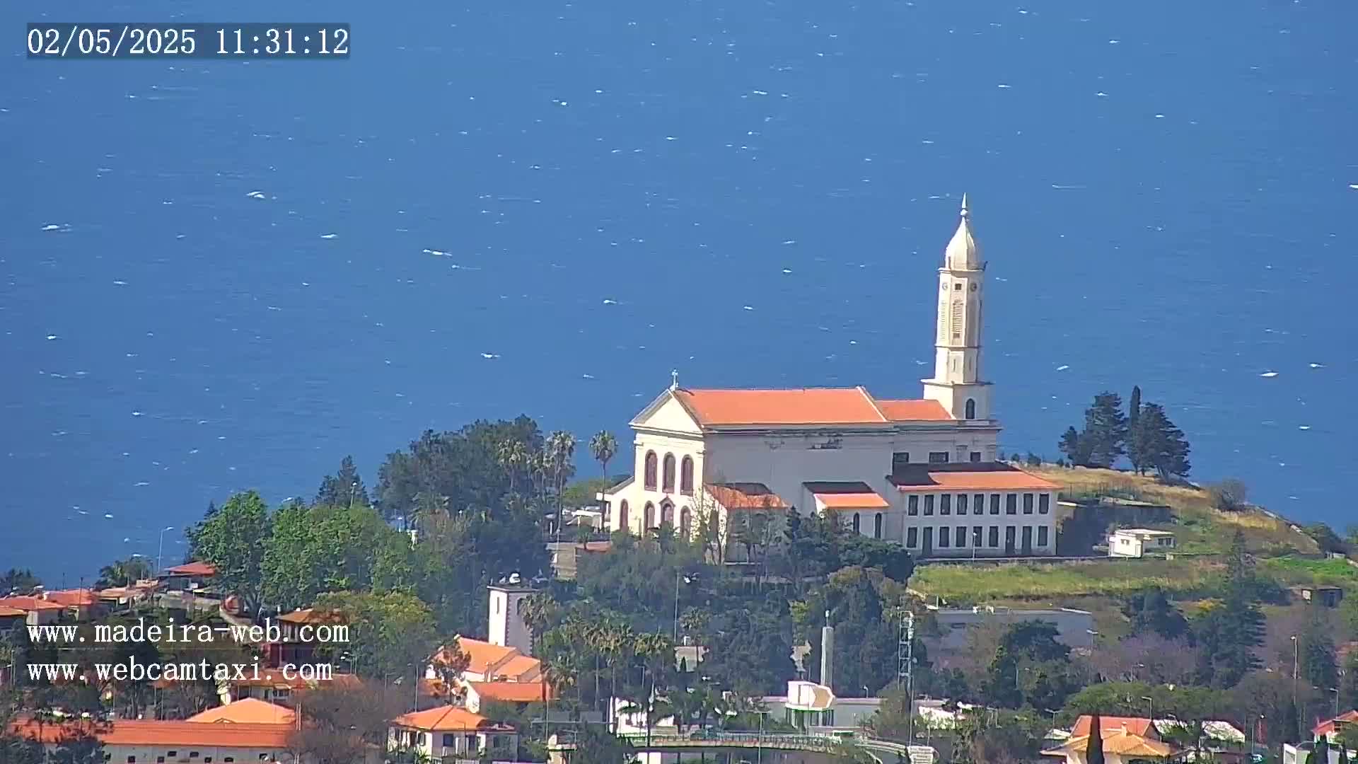 A white church with a tall steeple sits on a hill overlooking a blue ocean on a partly sunny day.