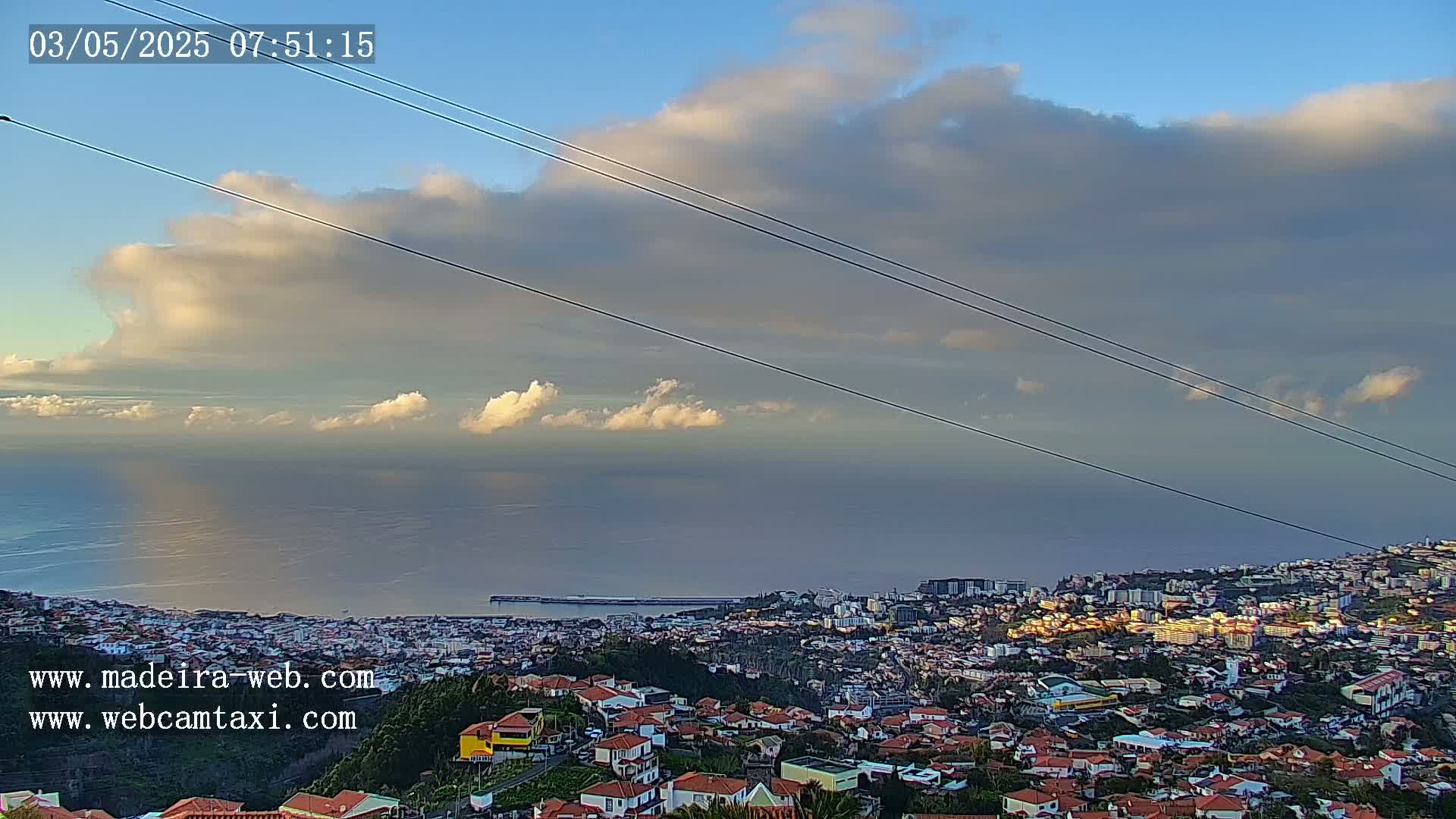 A coastal town sprawls beneath a partly cloudy sky, with the ocean stretching to the horizon under a mostly clear sky.