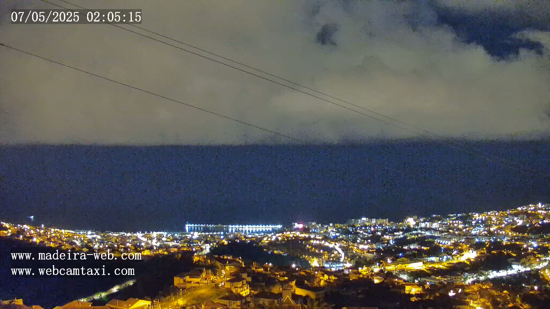 A nighttime view of a coastal city illuminated with lights, under a cloudy sky, overlooking a dark ocean.