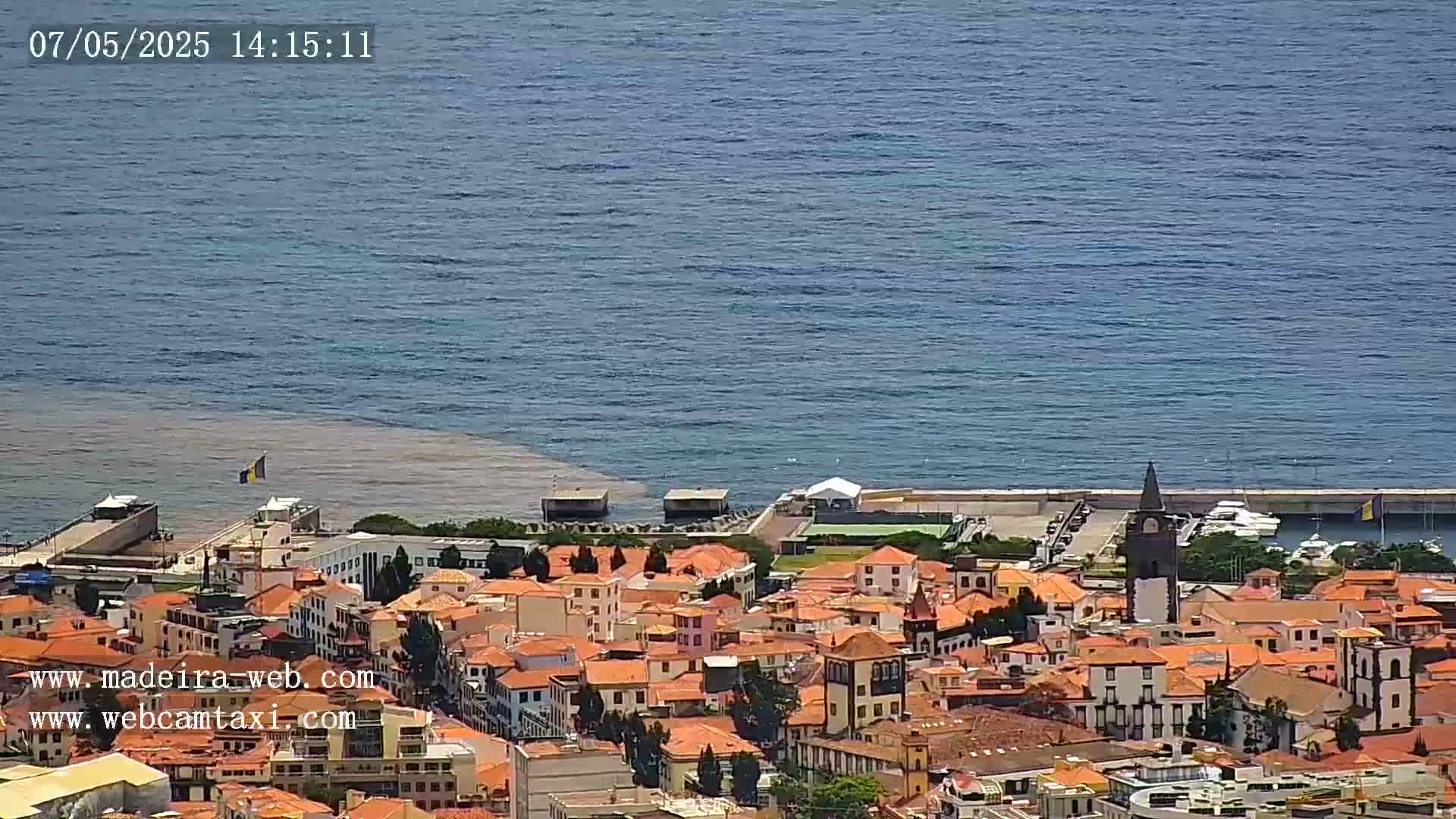 A coastal town with red-roofed buildings and a church steeple sits beside a calm blue ocean with a section of murky brown water near the shore.
