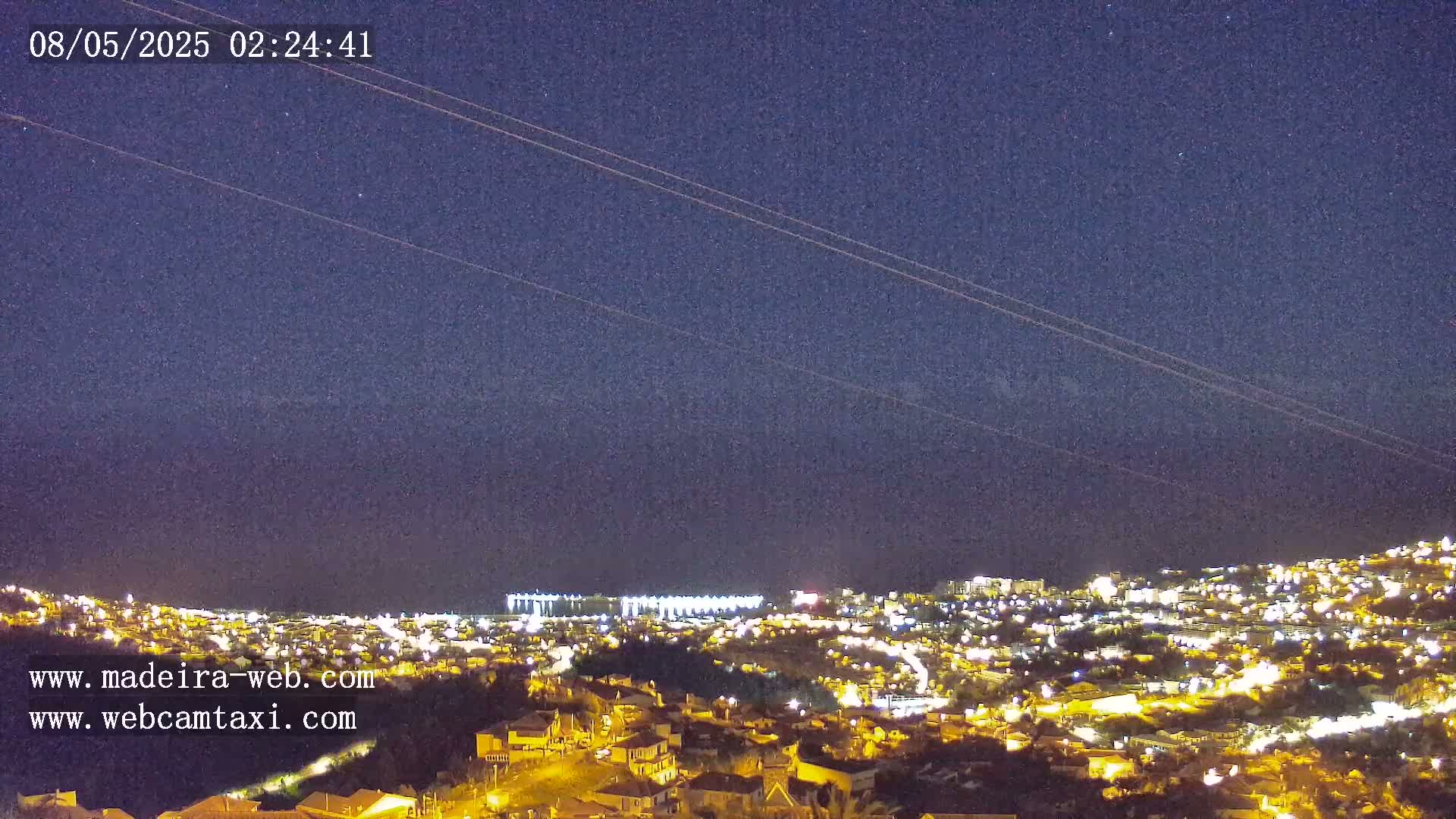 A nighttime view of a city illuminated by lights, stretching down a hillside to the ocean under a dark, clear sky.